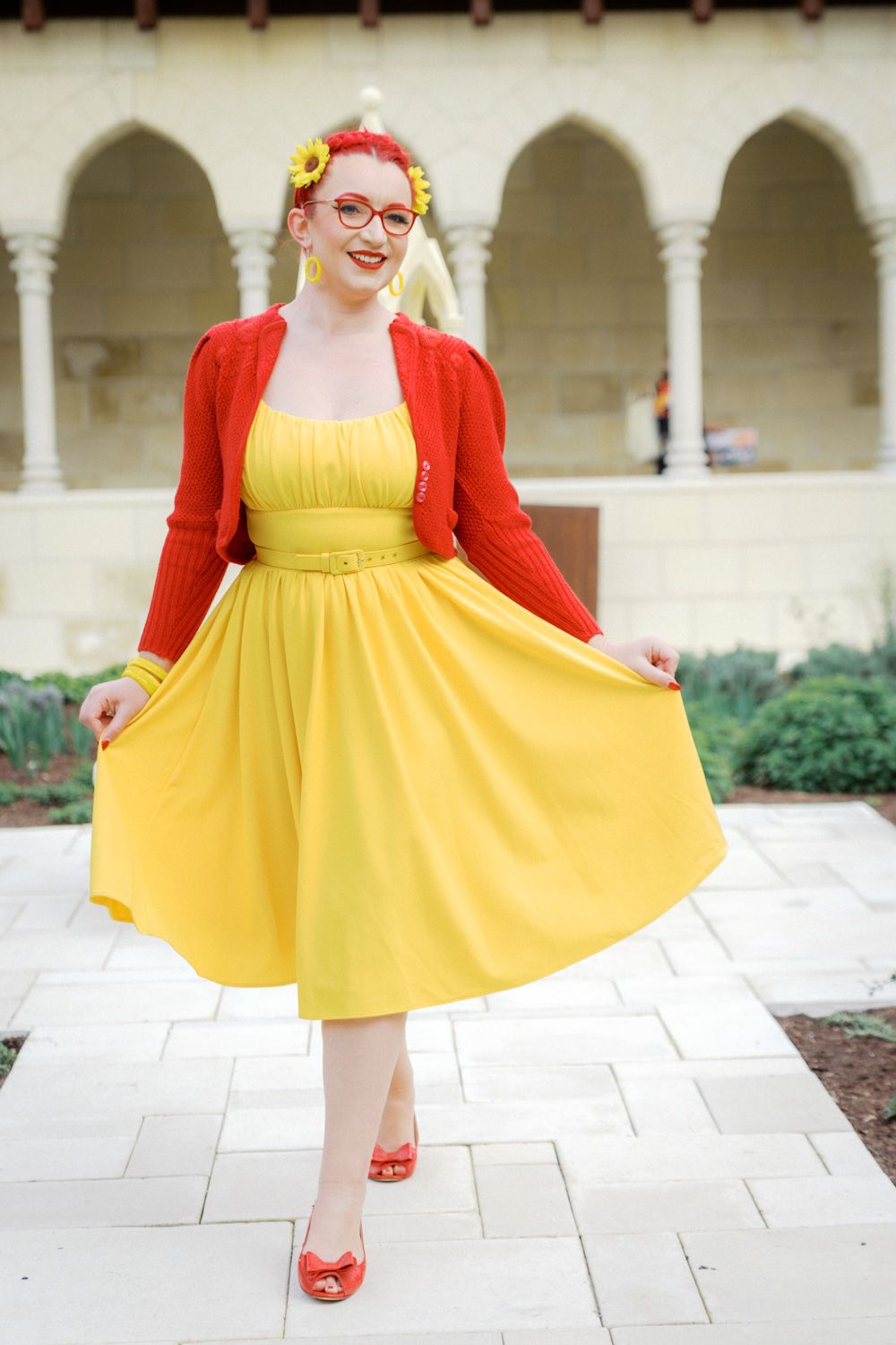 Woman in yellow midi dress and red cardigan twirling outdoors near arched building.