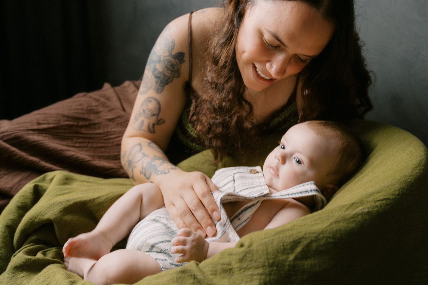 Tattooed mother smiling and bonding with her newborn baby on a green blanket.