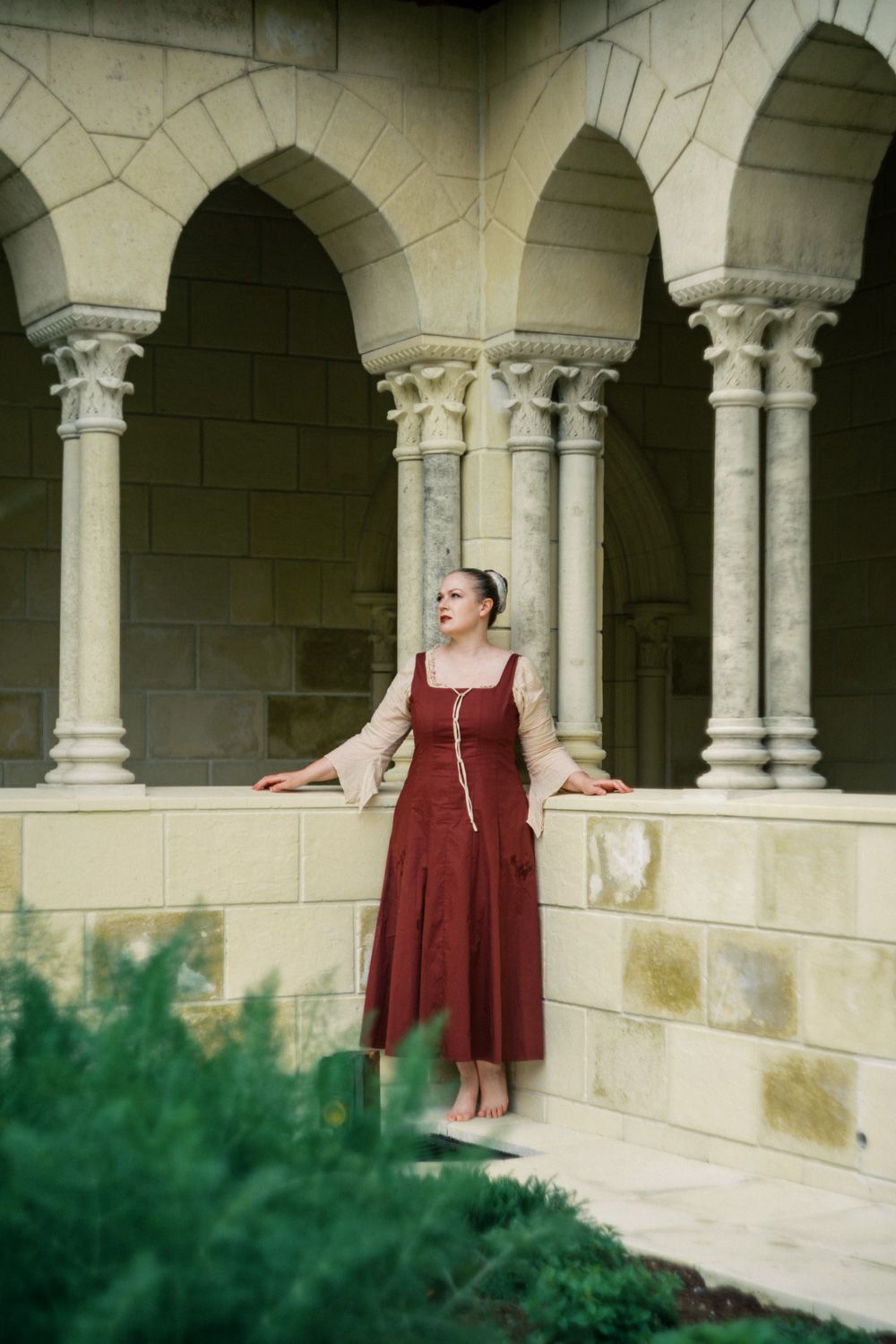 Woman in red medieval dress poses gracefully among stone arches and columns of a historic building.