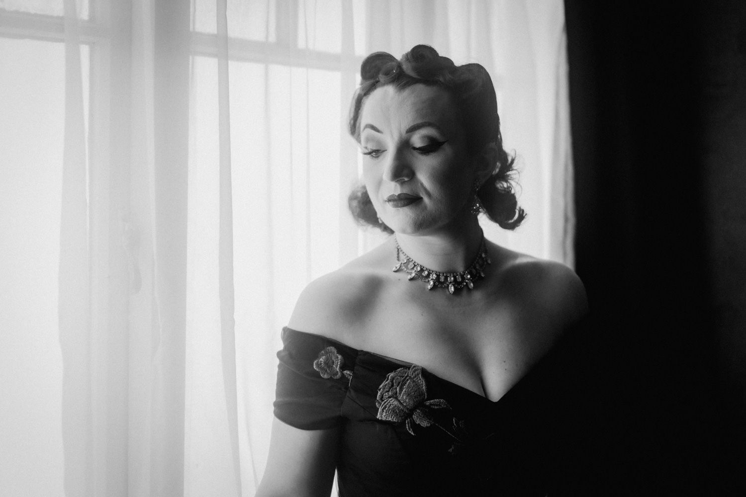 Black and white portrait of an elegant woman in off-shoulder gown with floral detail and diamond necklace by sheer curtains.