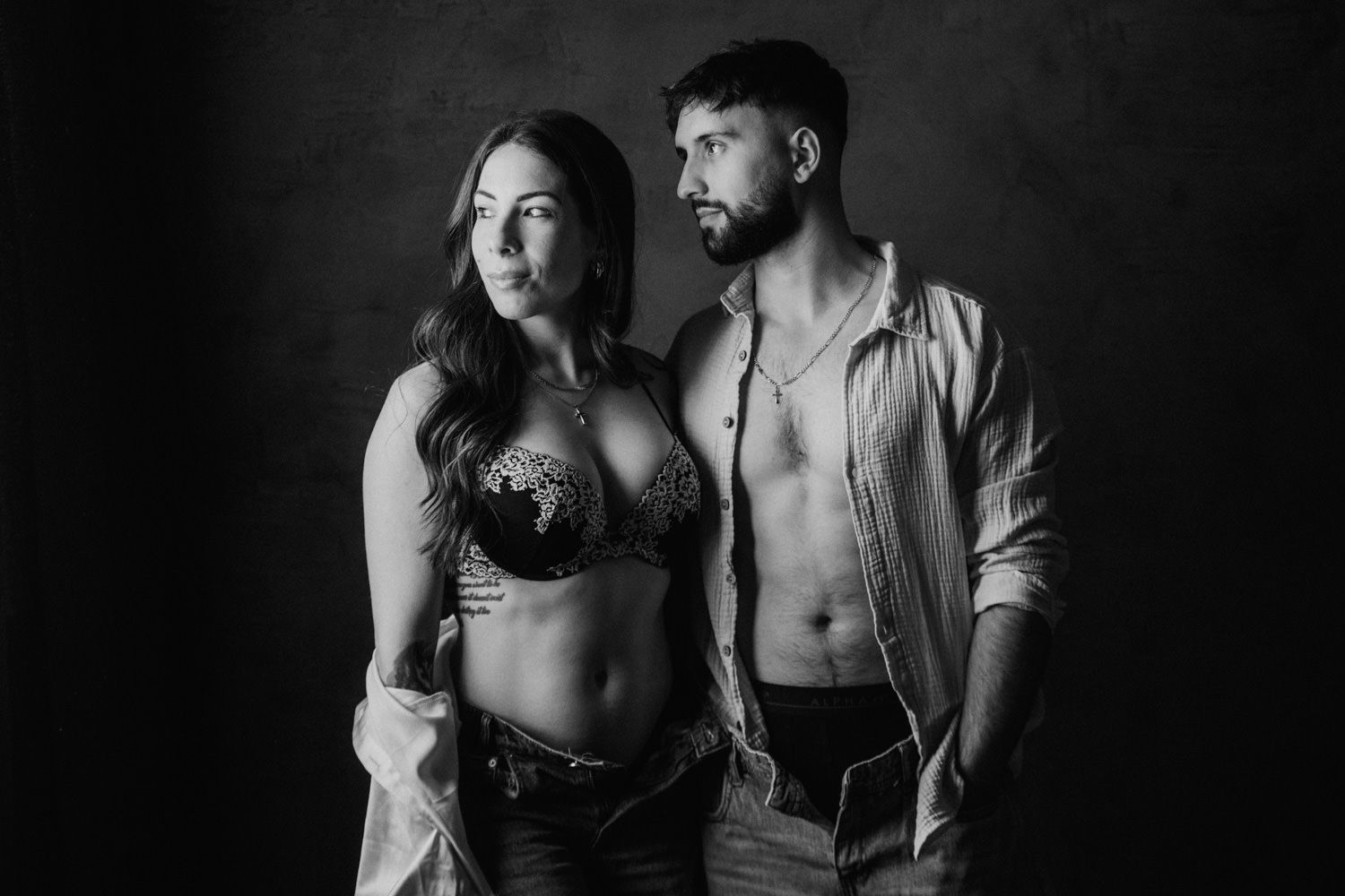 Black and white photo of a couple posing intimately, woman in lace bra and man in open shirt against dark background.