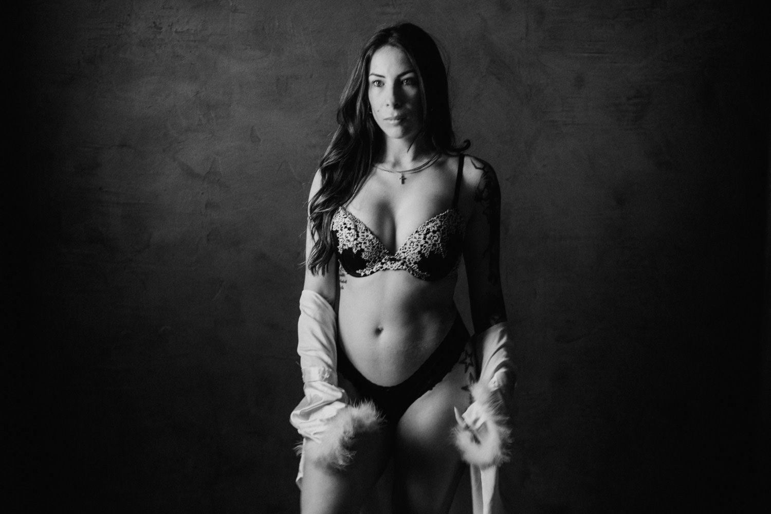 Black and white photo of a woman posing in lingerie and open robe against a dark studio background.