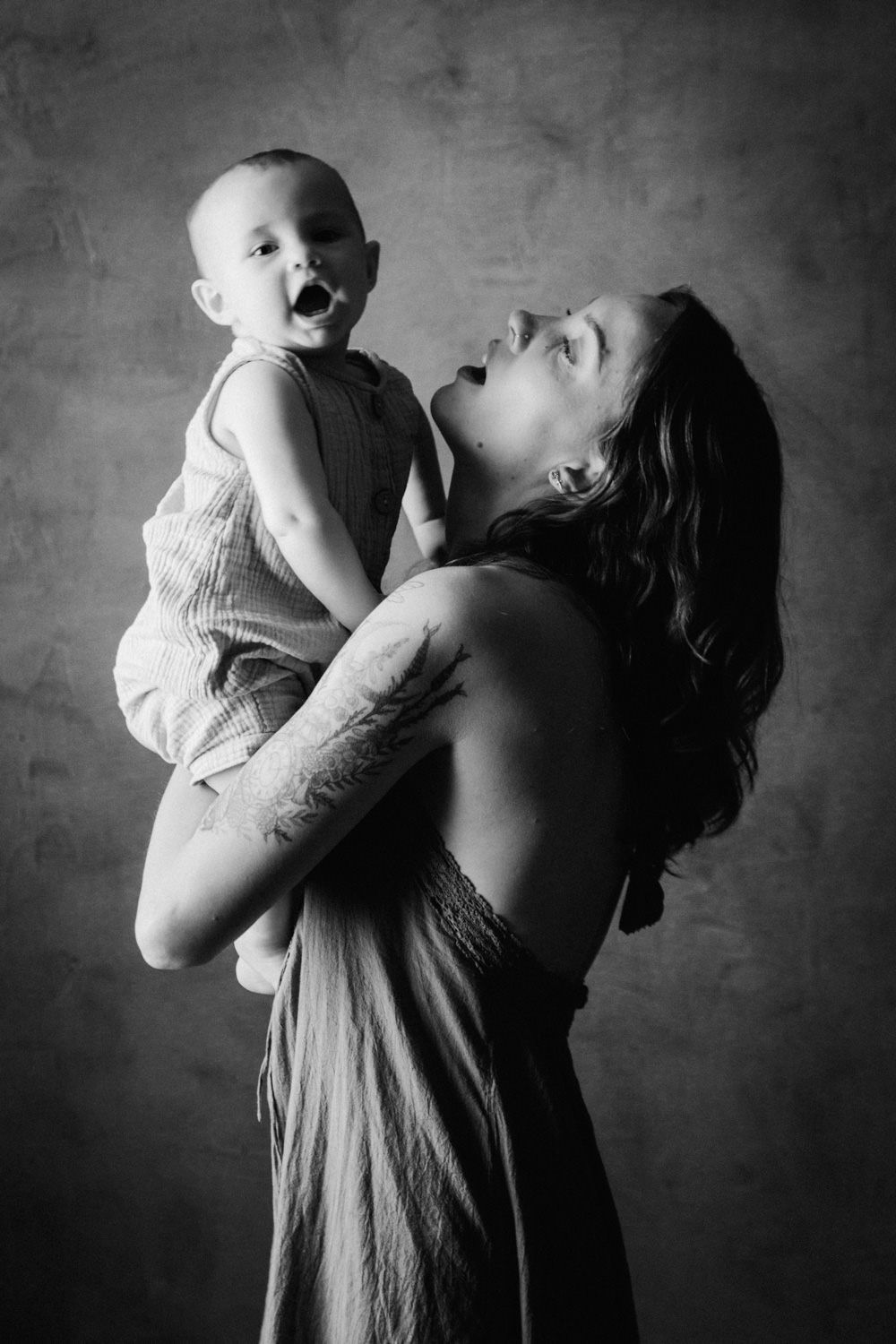 Black and white photo of a tattooed mother joyfully holding her laughing baby against a textured studio backdrop.
