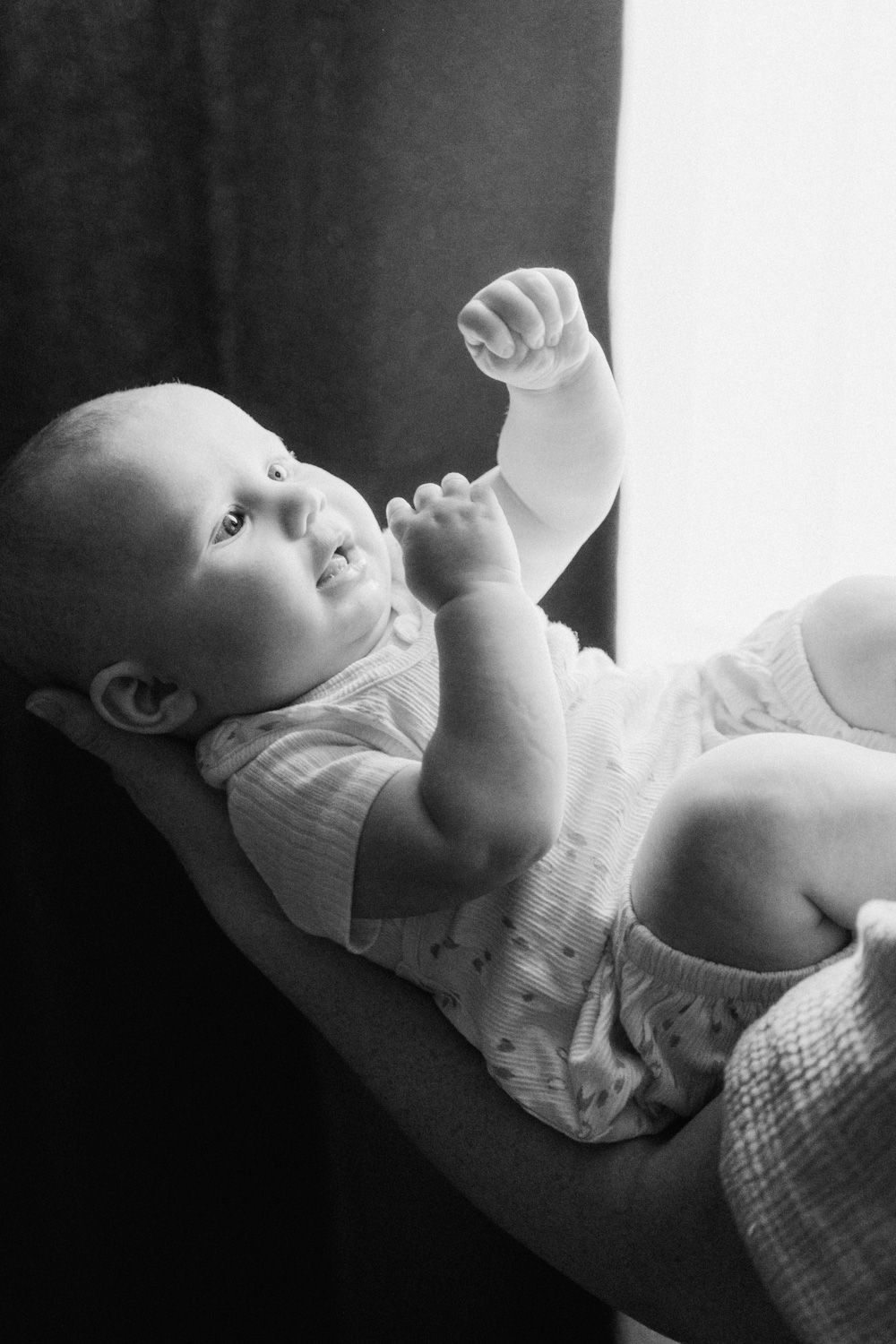 Black and white photo of a cute baby lying on their back, raising a tiny fist while looking upward with bright curious eyes.