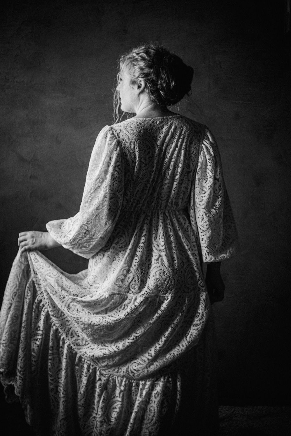 Black and white portrait of a woman in an elegant lace Victorian dress, viewed from behind against a dark backdrop.