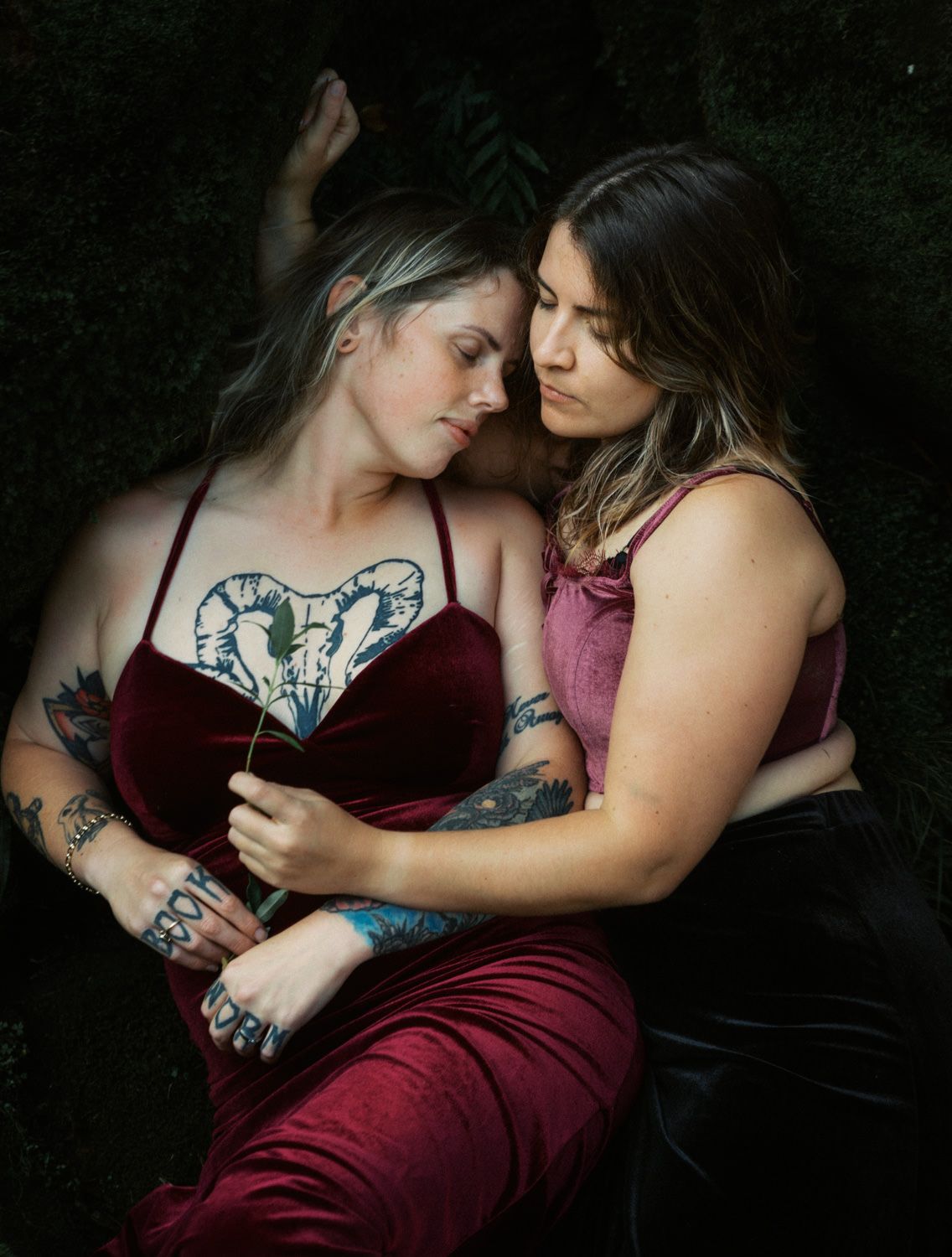 Two tattooed women in burgundy velvet outfits share an intimate moment, leaning close together outdoors.