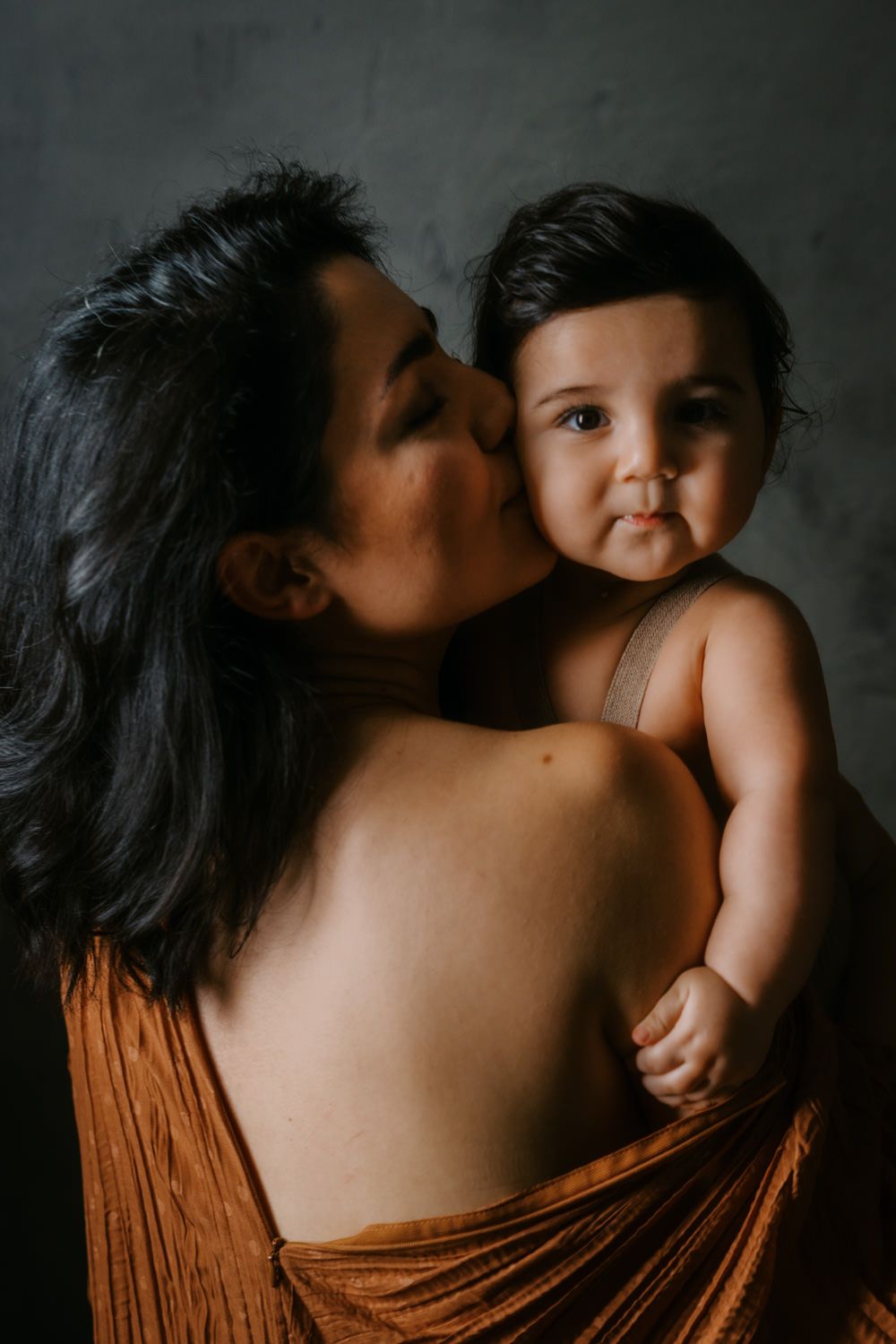 A mother with dark hair kisses her baby's cheek while holding the infant against her shoulder.