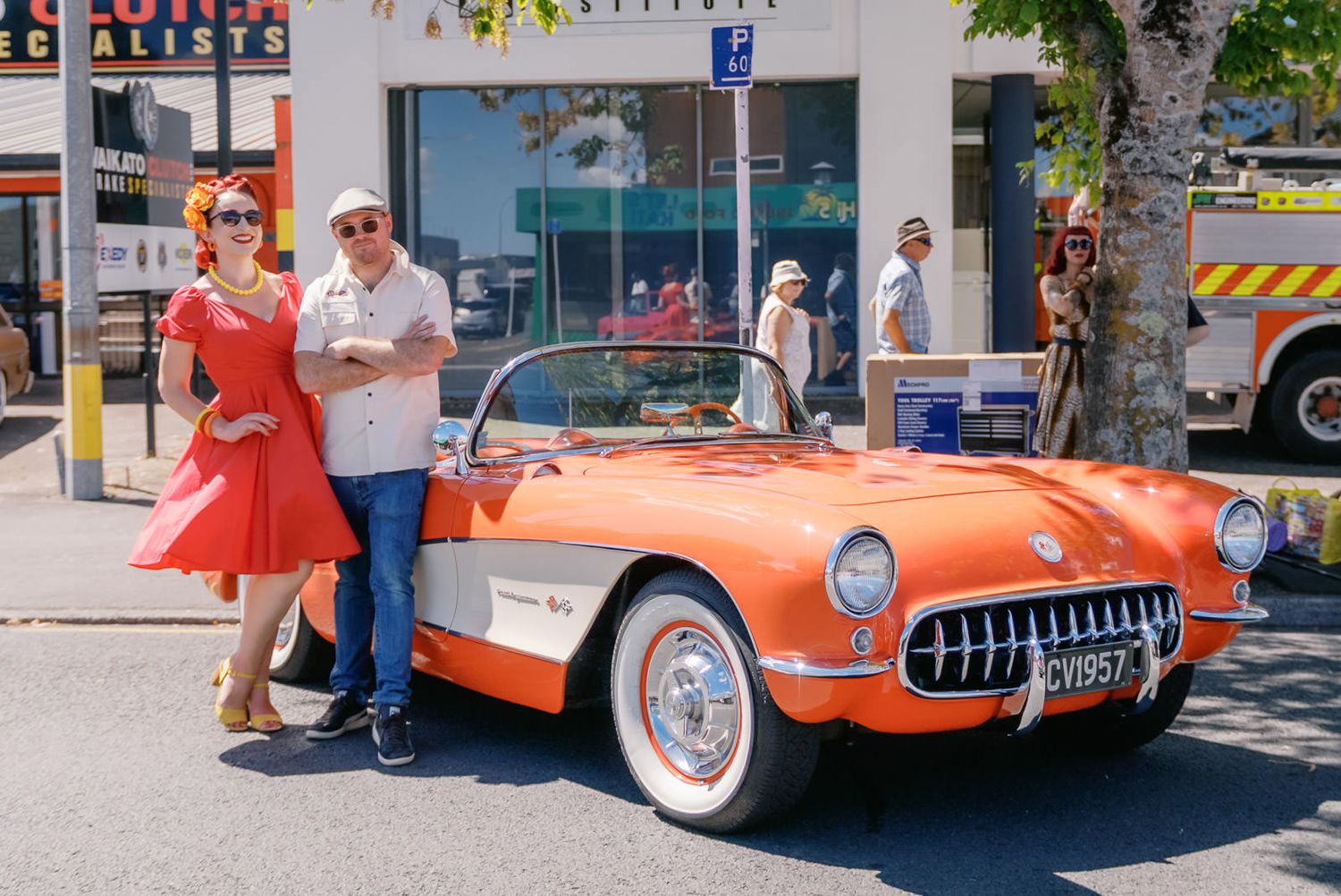 Couple posing beside a classic orange 1957 Corvette convertible at a sunny outdoor car show.