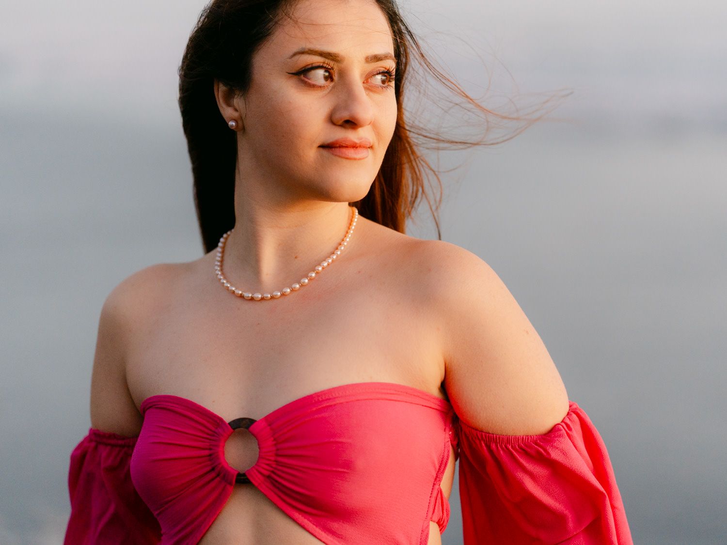 Young woman in pink off-shoulder crop top with pearl necklace posing outdoors against a cloudy sky.