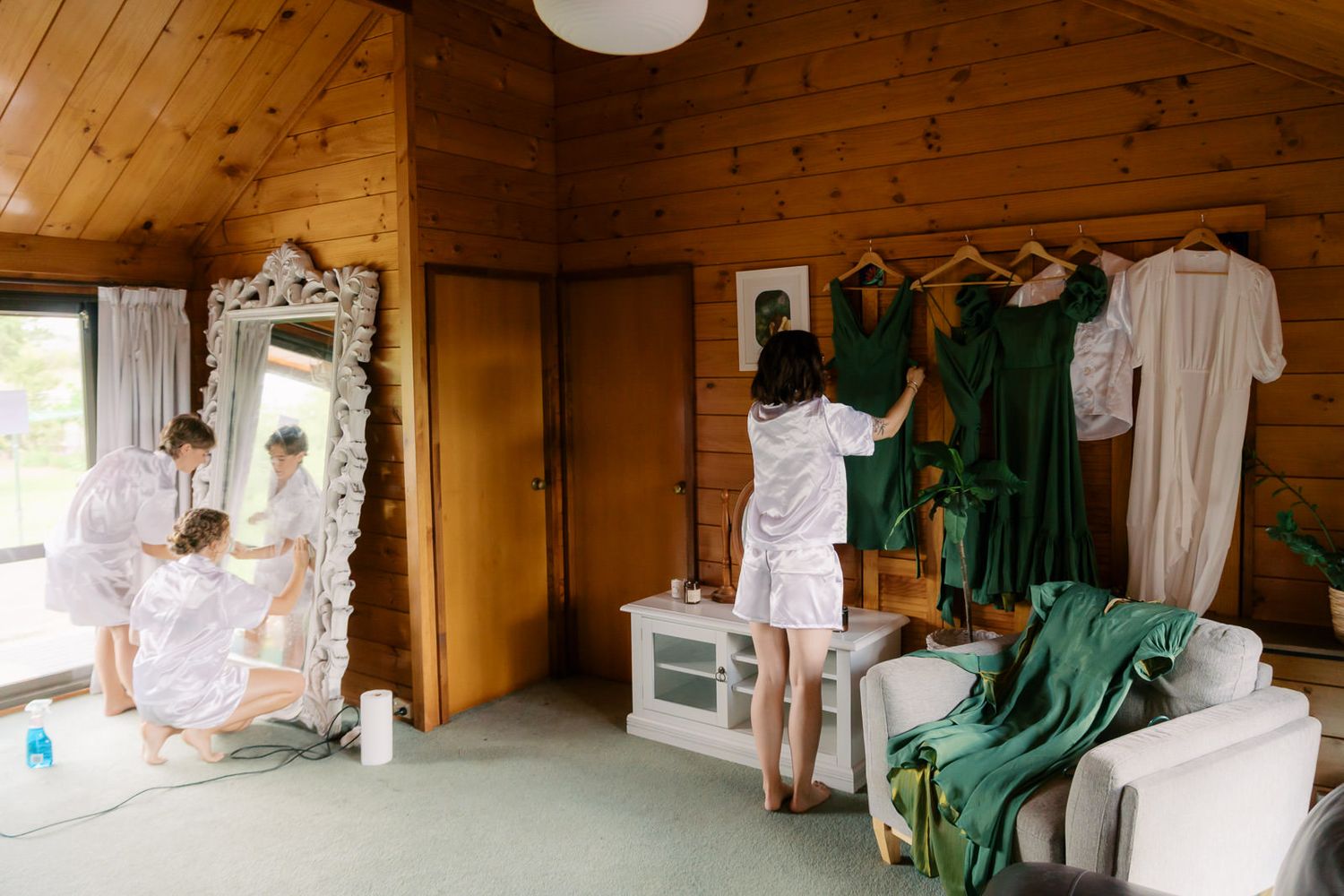 Bridesmaids in white robes preparing for a wedding, arranging green dresses in a rustic wood-paneled room with a ornate mirror.