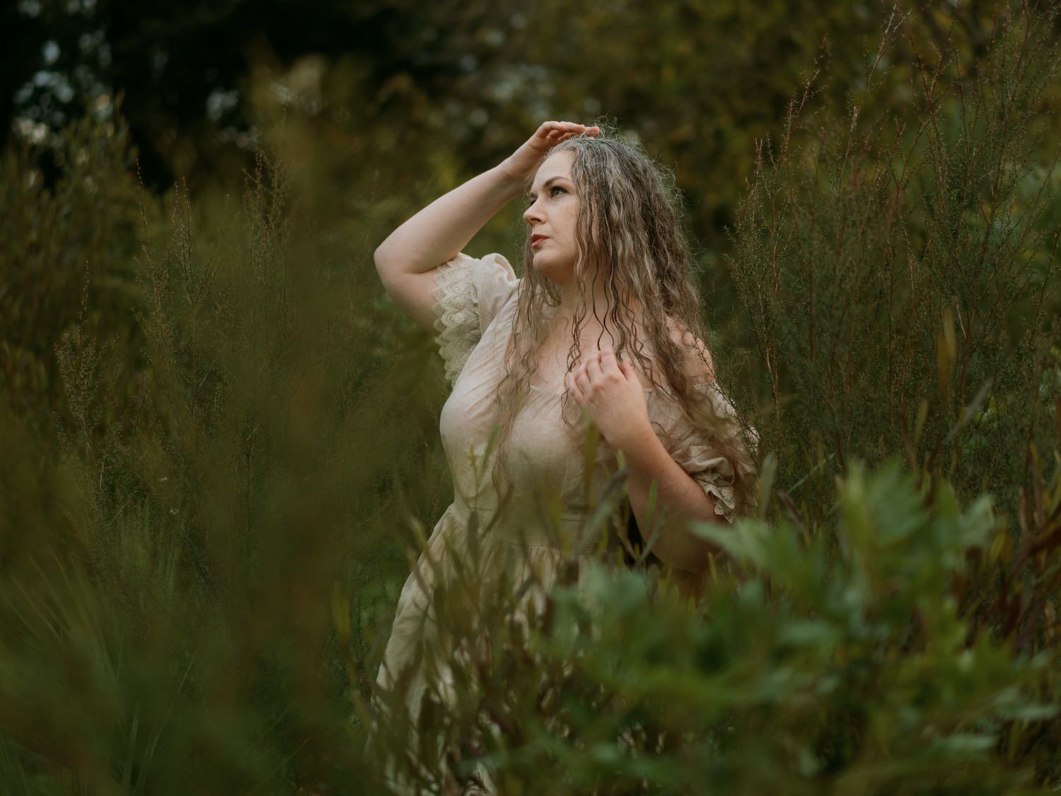 A woman with long wavy hair poses among lush green foliage in a dreamy outdoor nature setting.