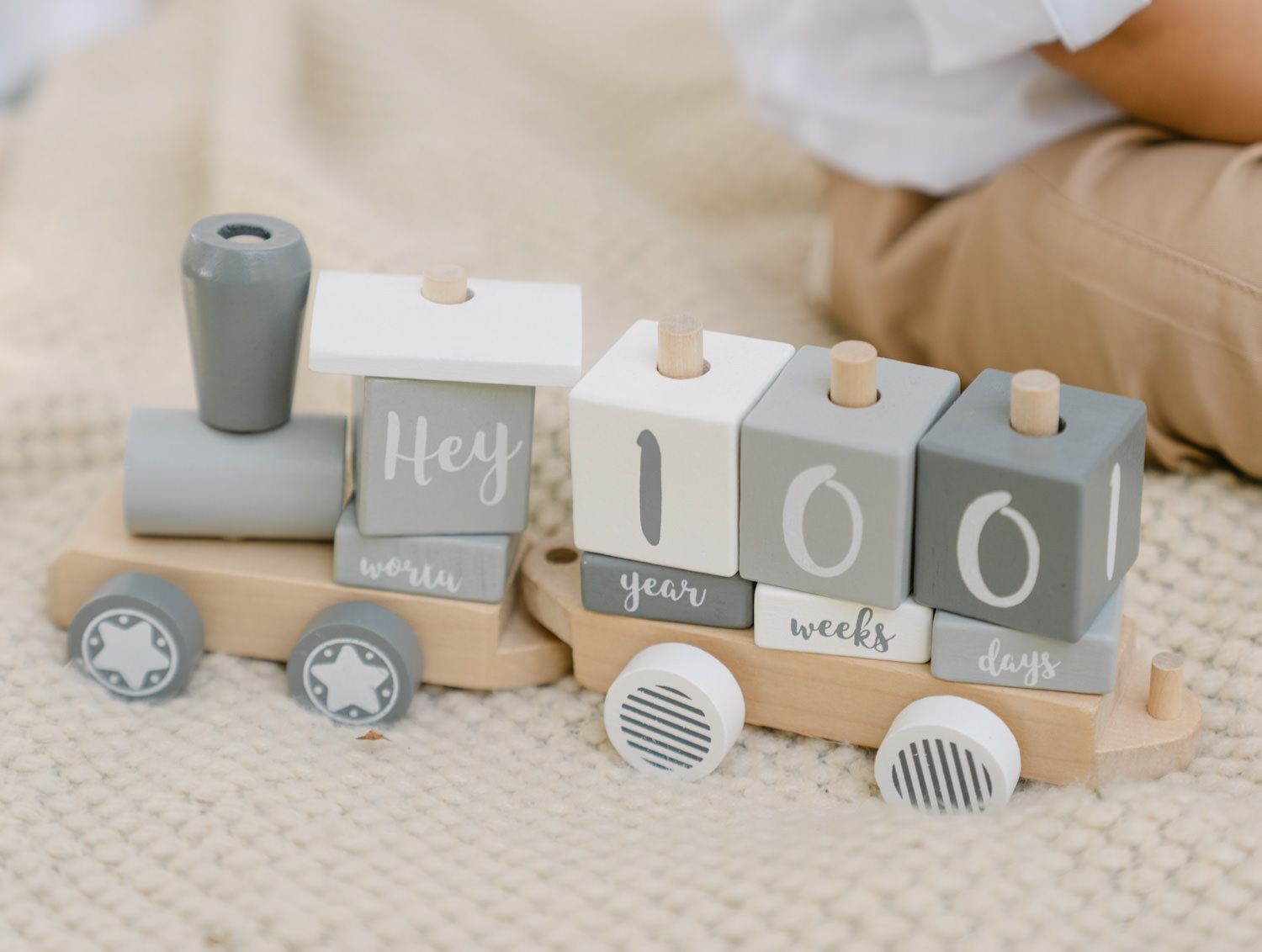 Wooden baby milestone train toy with gray and white blocks displaying Hey, 100, weeks, days, and world on a knit blanket.