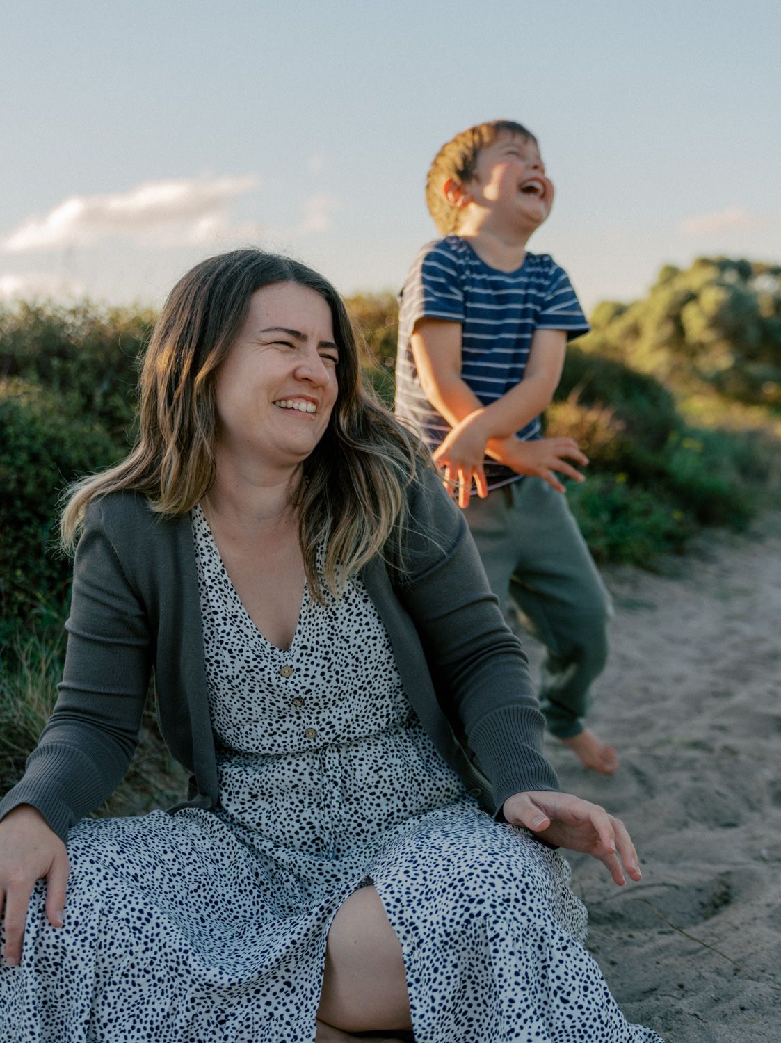 A woman in a floral dress sits on sandy ground laughing while a young boy in a striped shirt plays joyfully behind her.