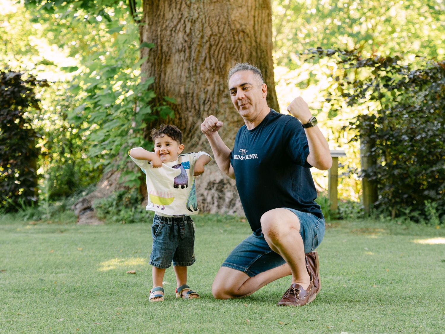 Man and toddler boy flexing muscles together on green grass in a sunny park with large tree backdrop.