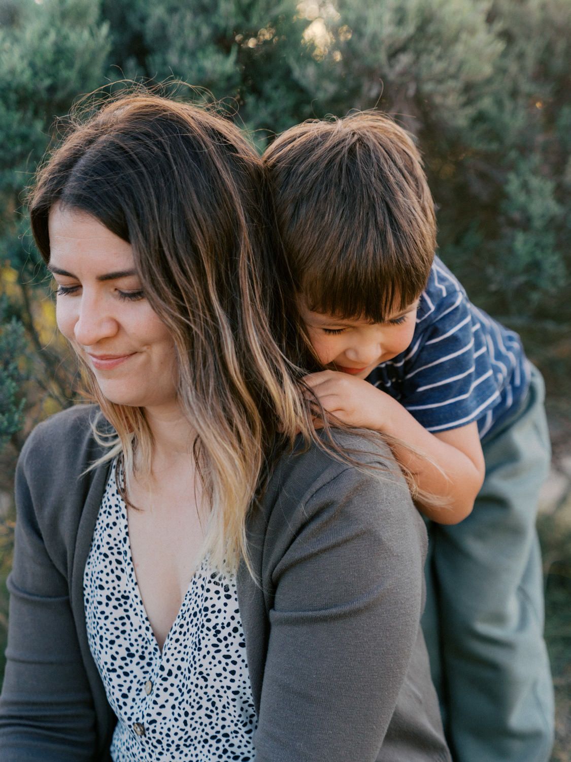 A mother smiles softly while her young son rests on her back outdoors among green trees.