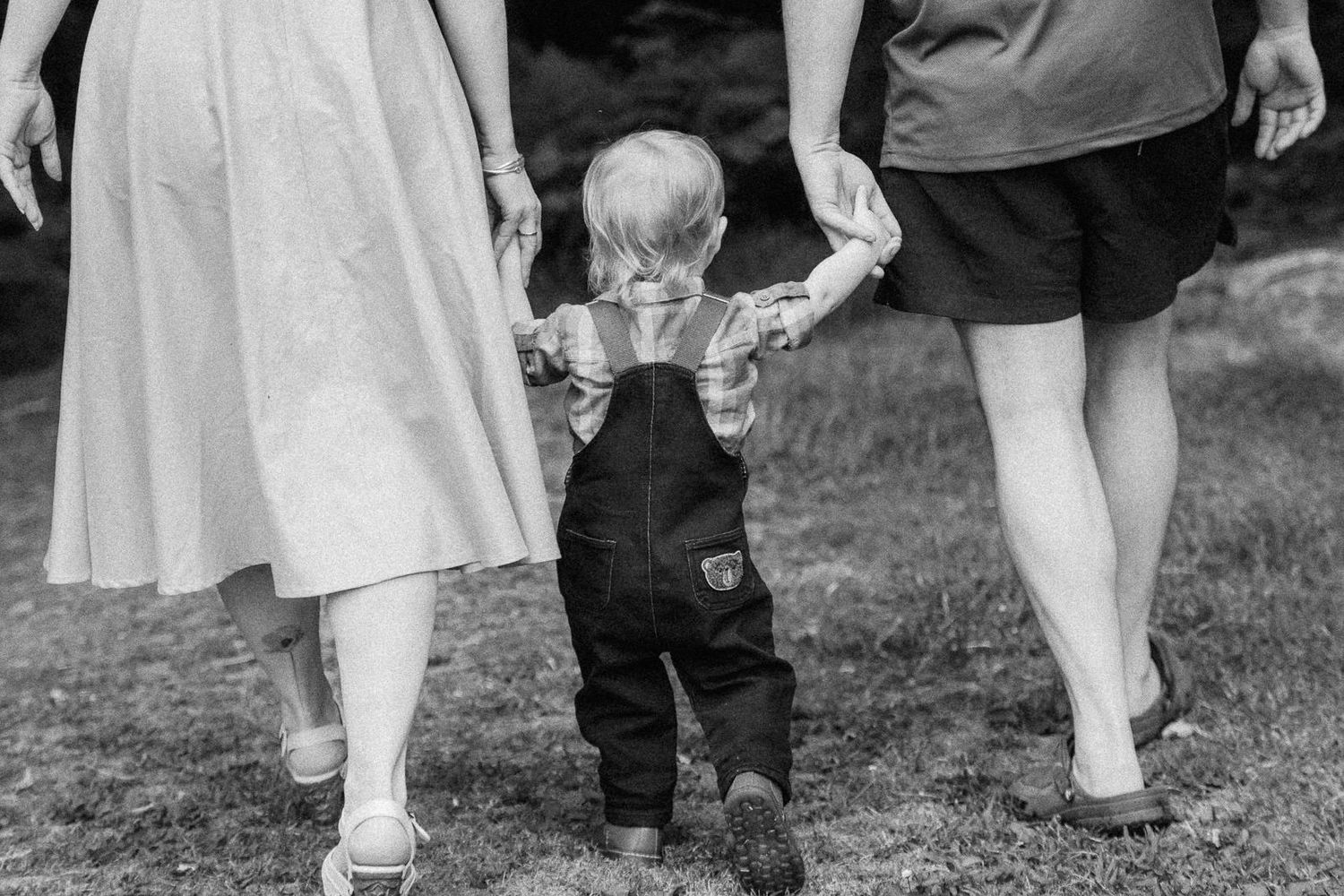 Black and white photo of toddler in overalls holding hands with two adults walking outdoors.