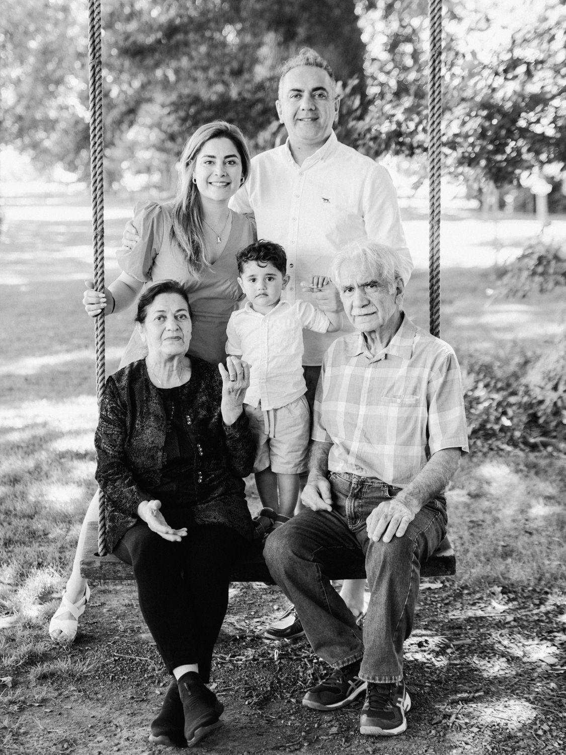 Black and white family portrait of five people posing on a swing in a park setting.