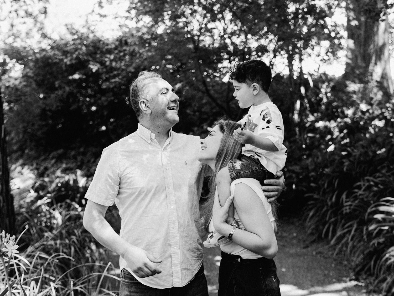 Black and white photo of a happy family with a man, woman, and young child outdoors in a lush garden setting.