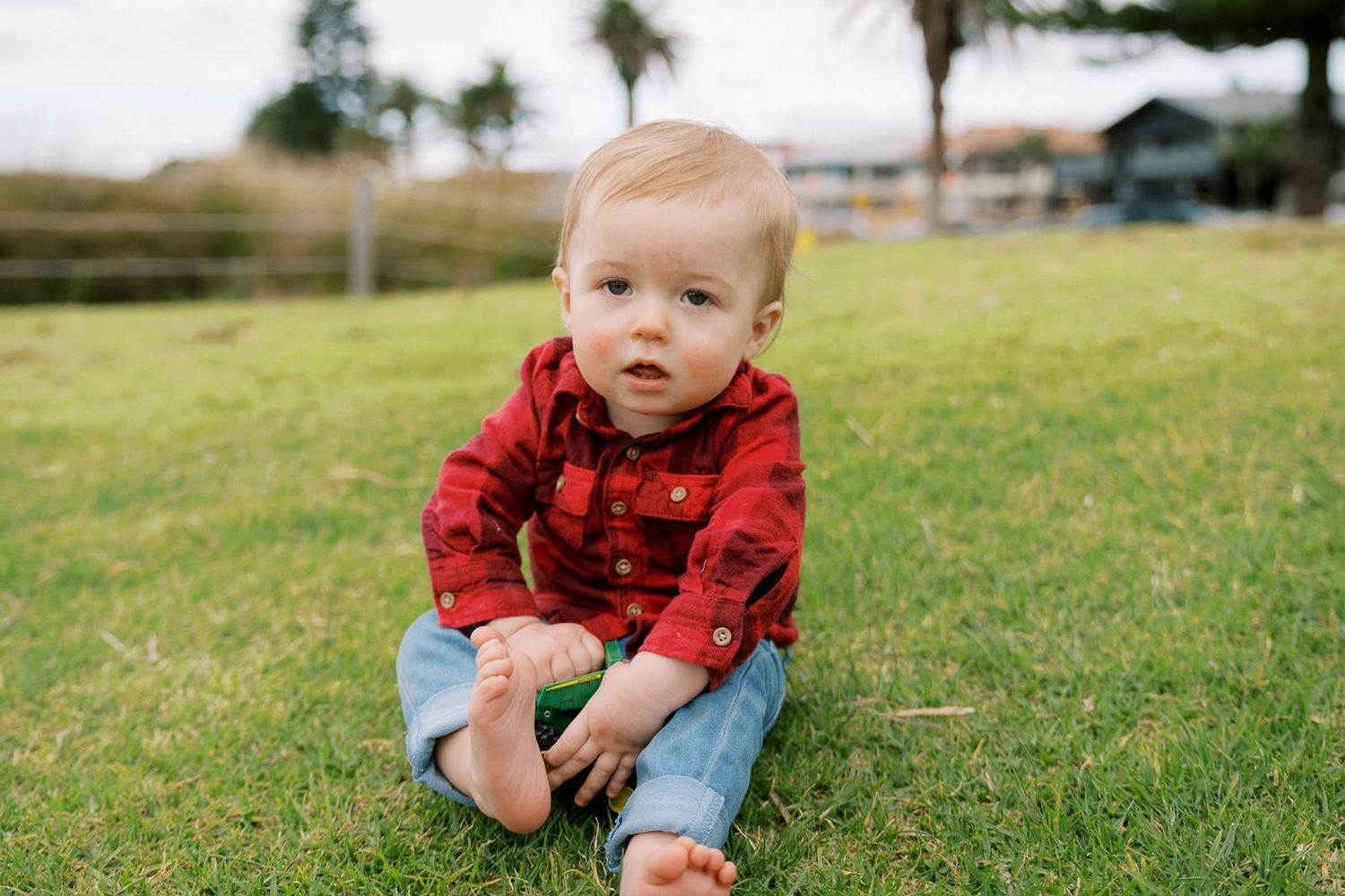 A cute baby in a red flannel shirt and jeans sits barefoot on green grass outdoors.