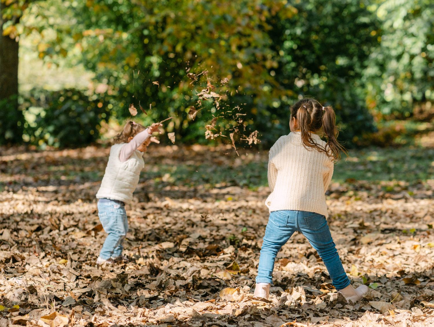 Two children tossing autumn leaves in a park, wearing white sweaters and jeans, surrounded by golden fallen leaves.