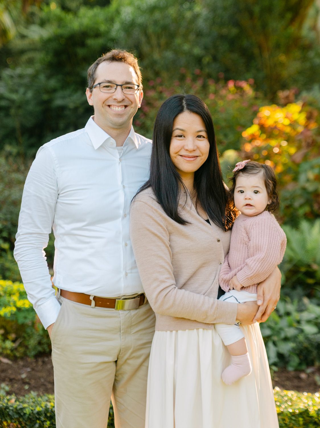 Young family of three with parents holding baby girl smiling in a colorful garden.