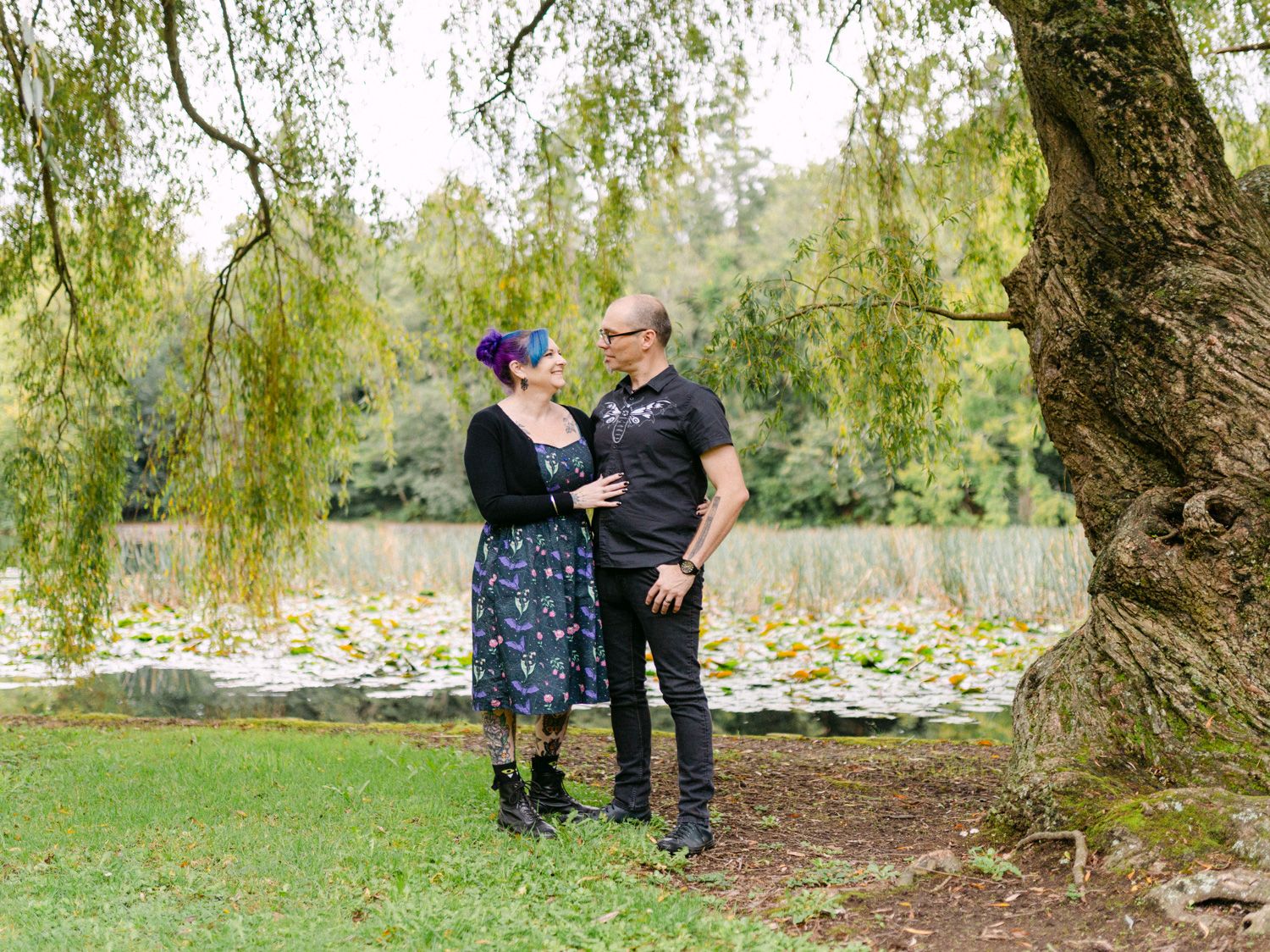 Couple standing under a willow tree by a pond in a lush green park, sharing a romantic moment.