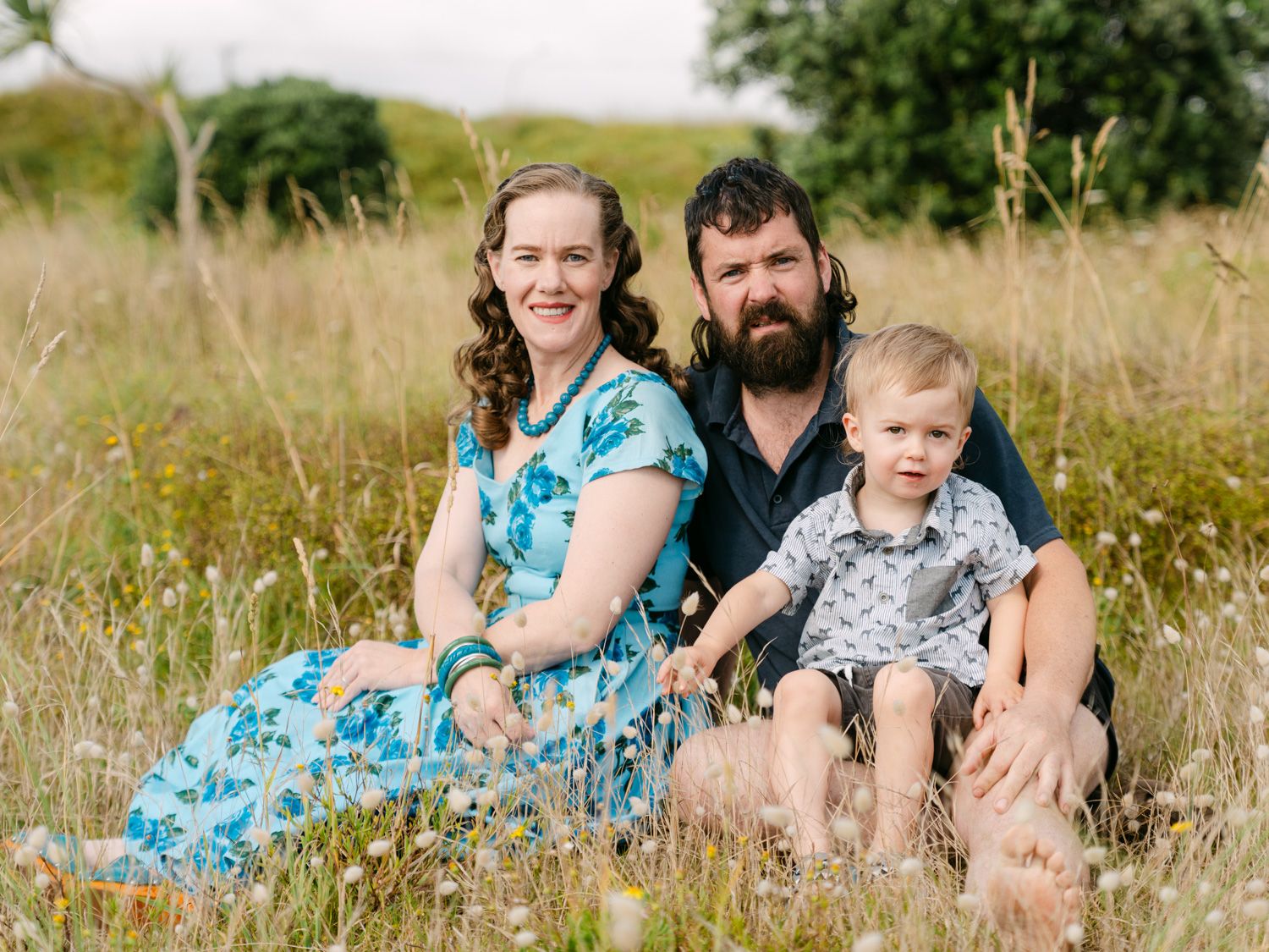 Family of three sitting in a grassy meadow, parents with young toddler smiling outdoors.