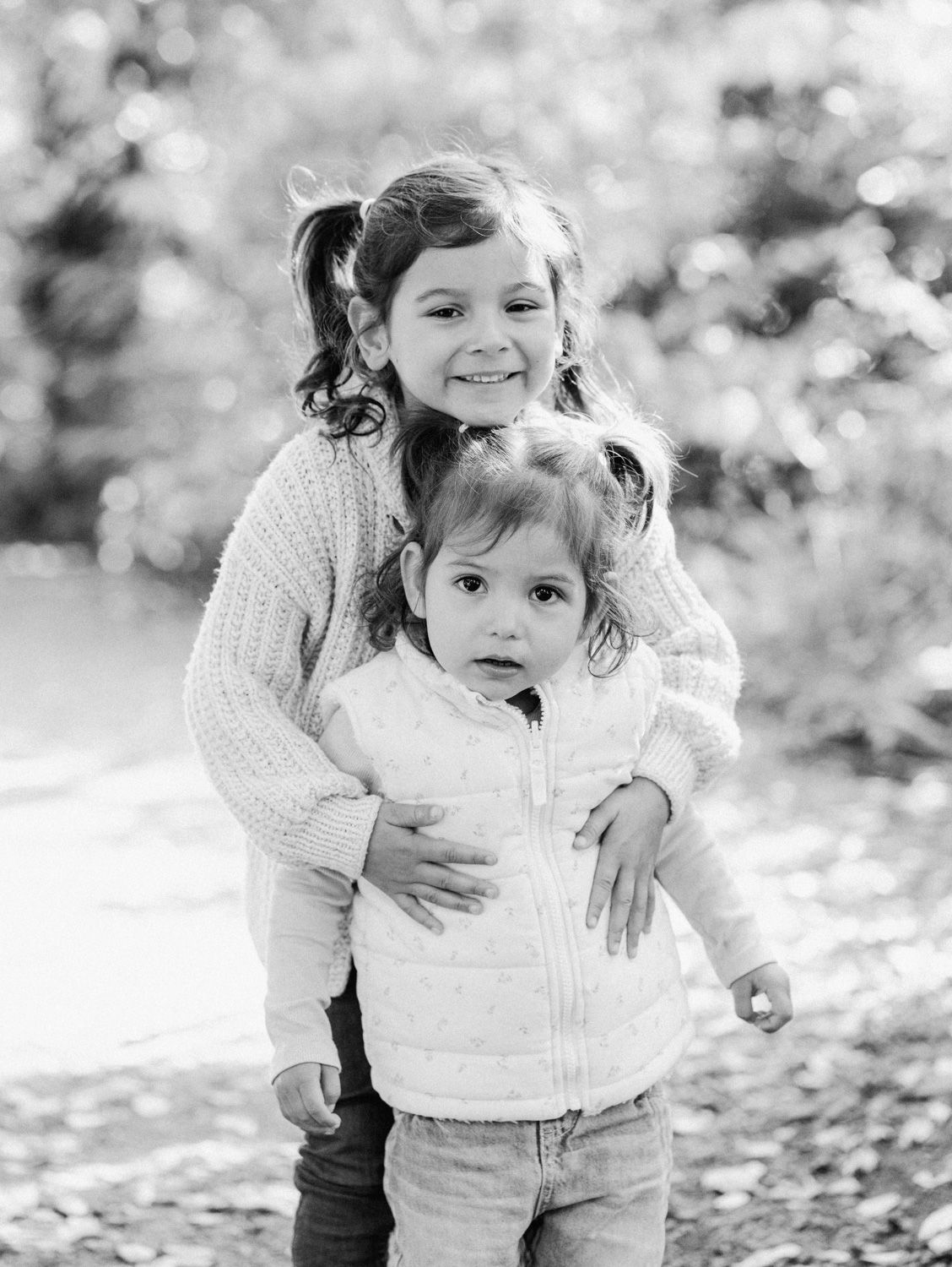Black and white photo of two young sisters hugging outdoors, older girl smiling with pigtails embracing toddler sibling.