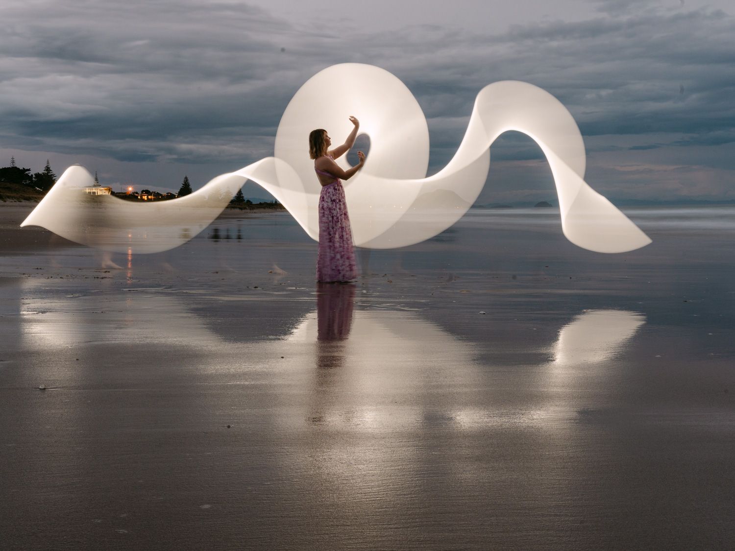 Woman in floral dress creates glowing light painting waves on reflective beach at dusk.
