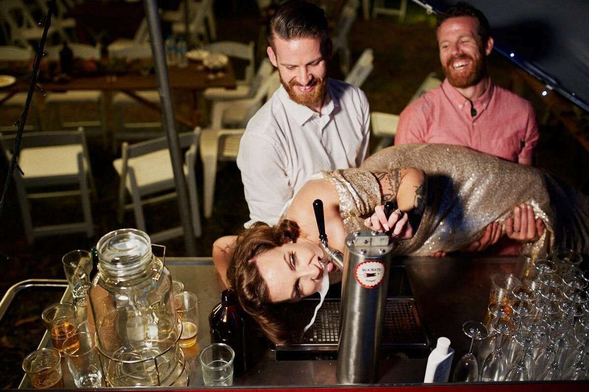 Two men laugh as a woman leans back over a bar surrounded by mason jar glasses at an outdoor evening event.