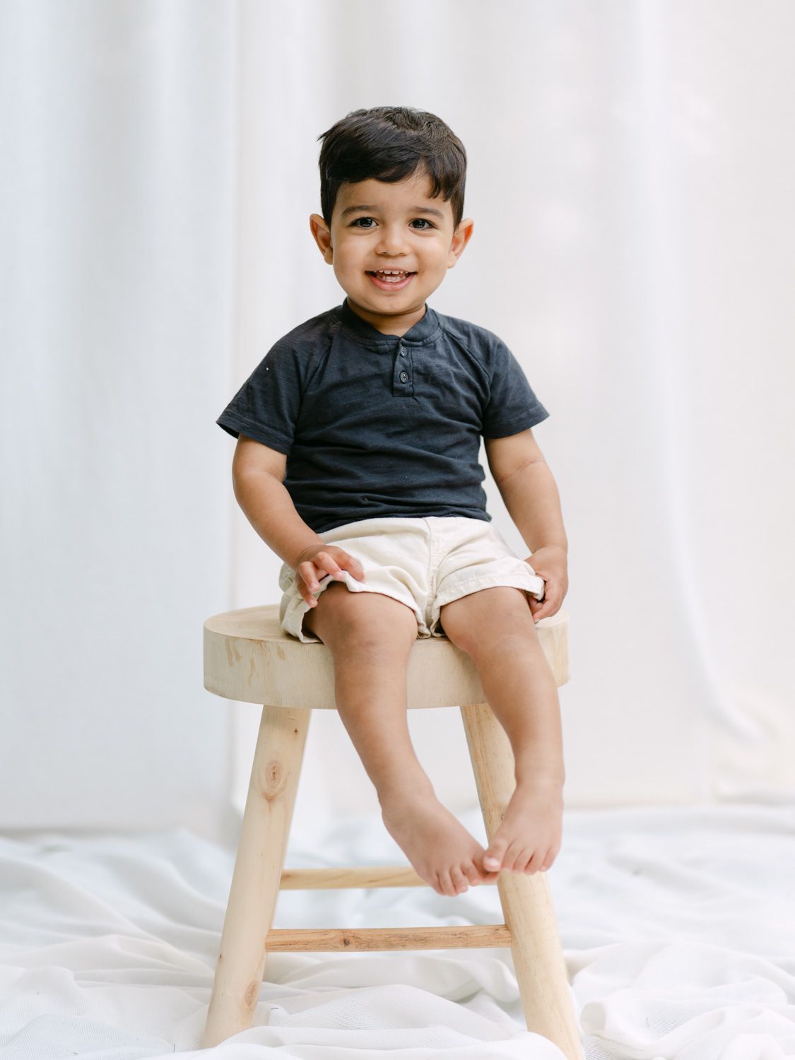 Smiling toddler boy in dark henley shirt and cream shorts sitting on a small wooden stool against white backdrop.