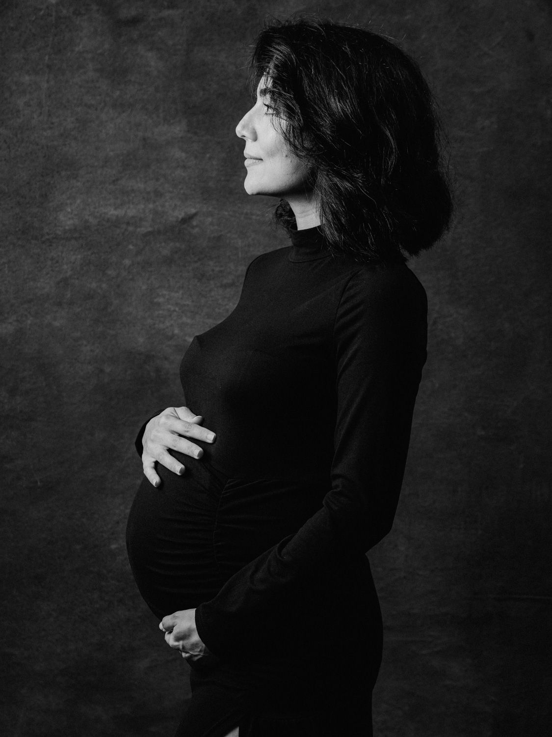 Black and white maternity portrait of a woman in a black turtleneck dress cradling her baby bump in a studio.