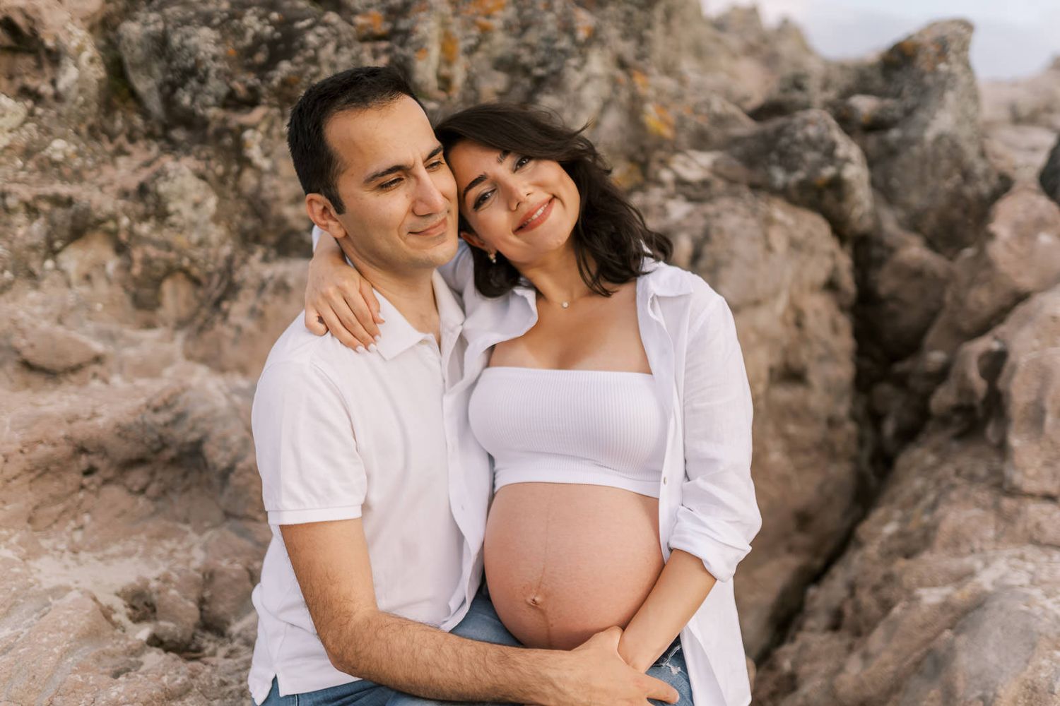 Couple posing outdoors against rocky backdrop, pregnant woman in white crop top cradling baby bump with partner embracing her.