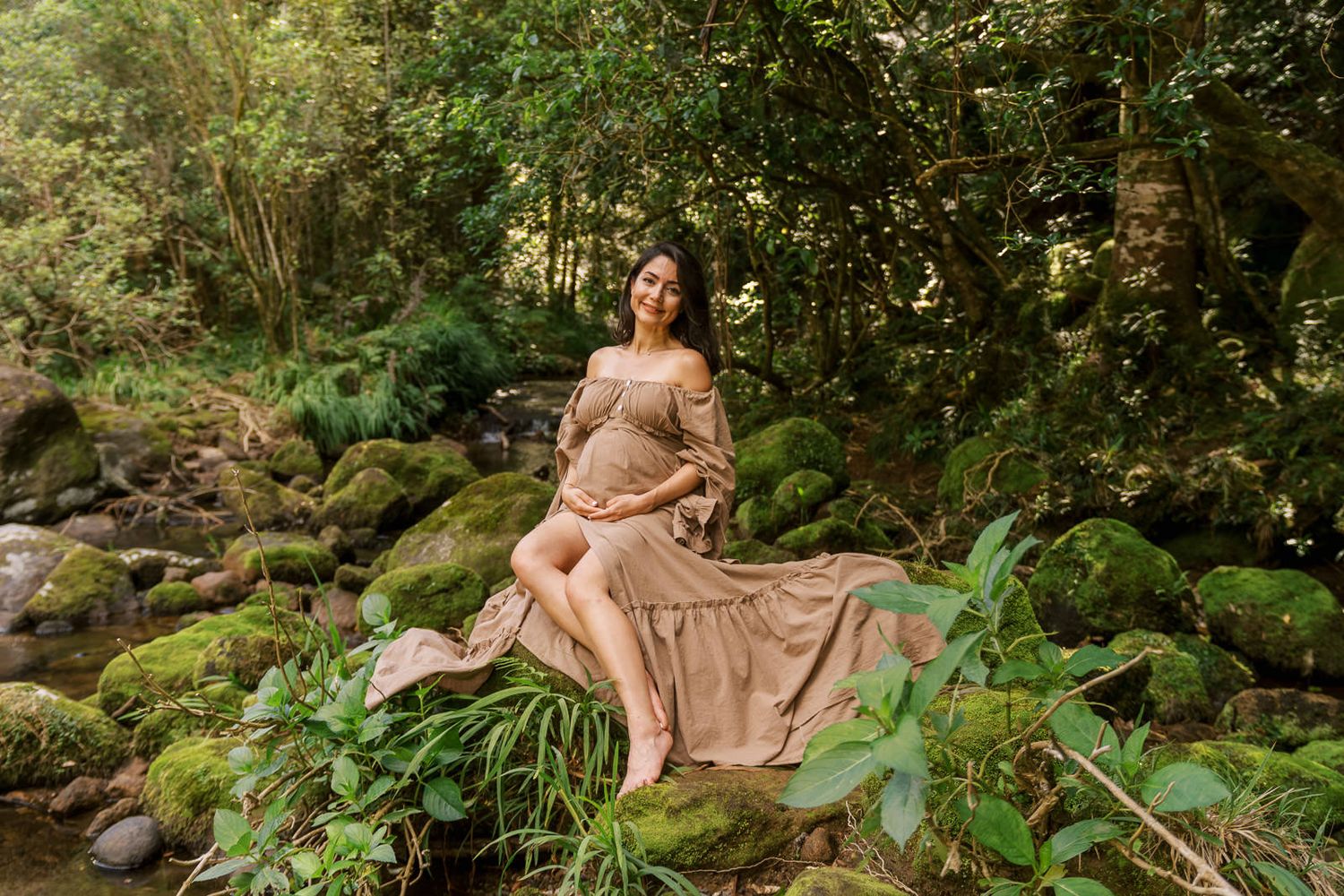 Pregnant woman in beige off-shoulder dress sitting on moss-covered rocks in a lush green forest setting.