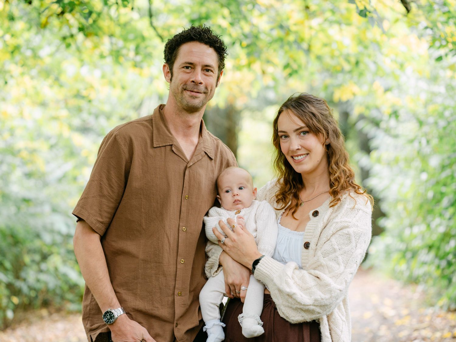 A young family with a newborn baby poses outdoors surrounded by lush green trees on a sunlit path.