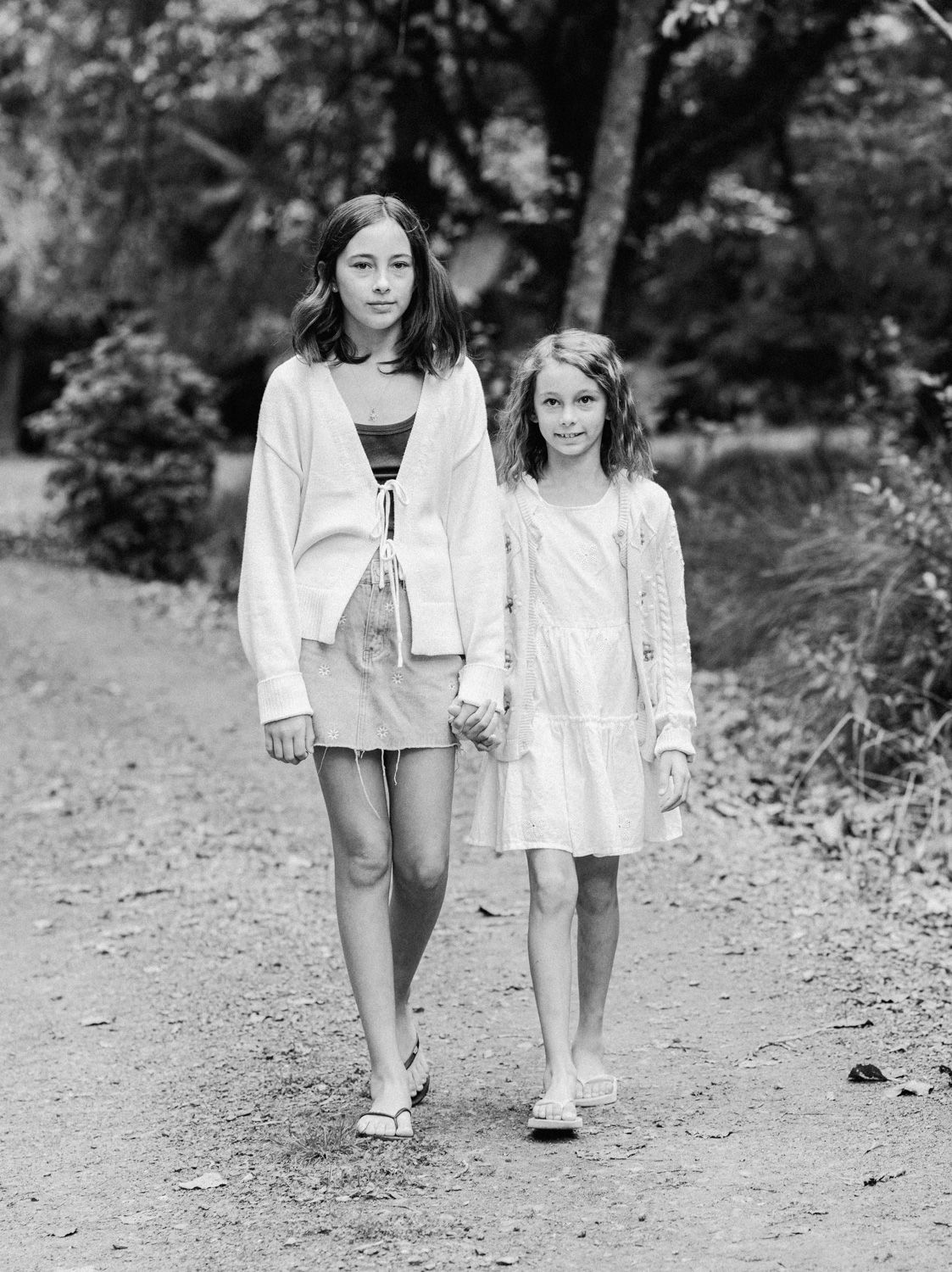 Two girls holding hands walk down a tree-lined path in a black and white portrait photo.