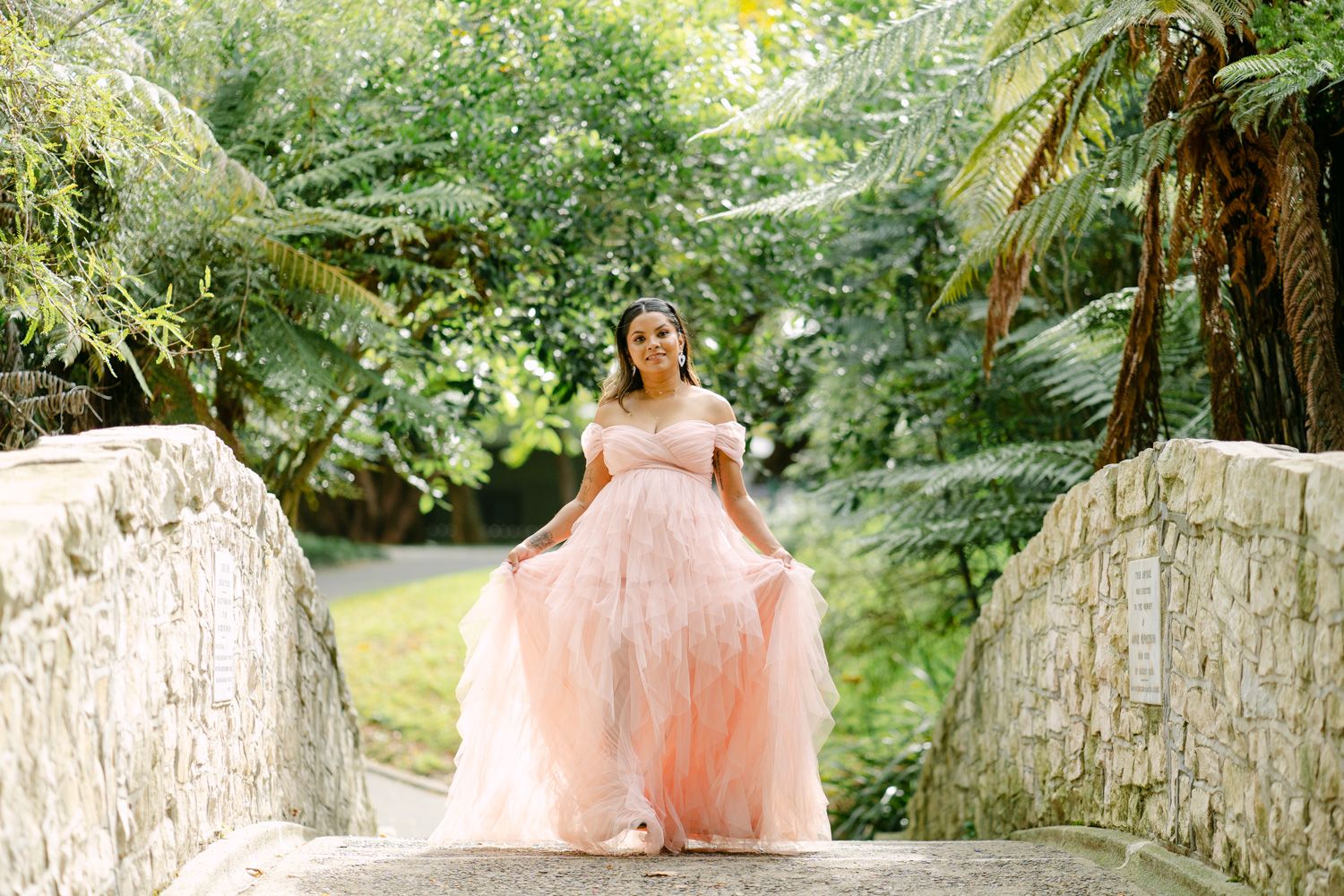 Woman in pink off-shoulder ball gown poses on stone bridge surrounded by lush tropical greenery.