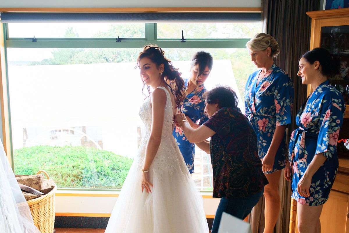 Bridesmaids in blue robes helping bride into her white wedding gown near a large window.