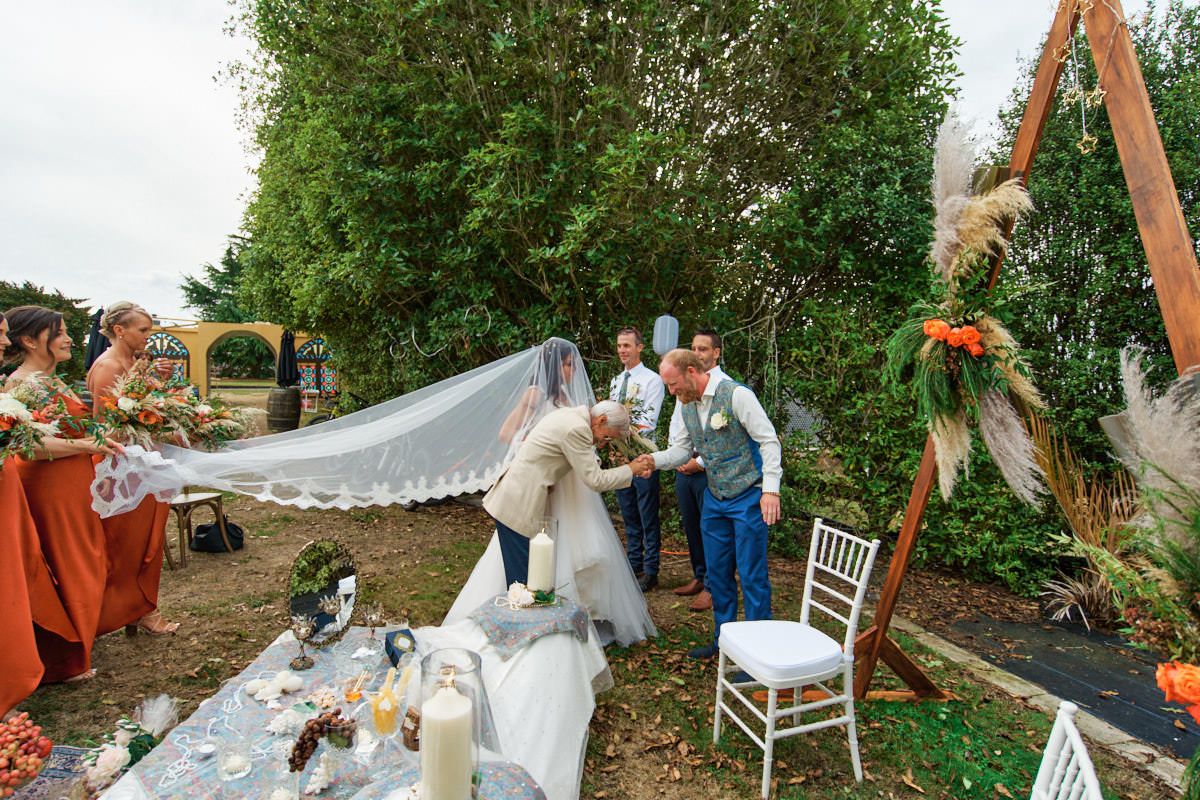 Bride and groom exchange vows at an outdoor wedding ceremony with orange floral decor and flowing veil.