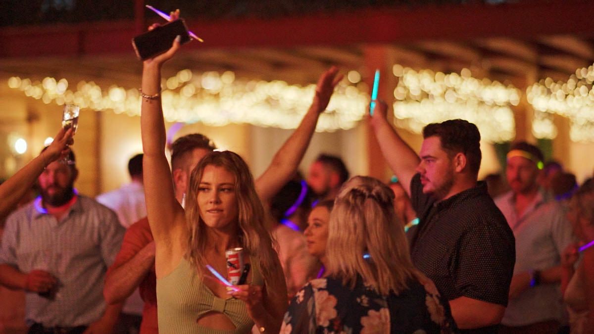 People dancing at an outdoor evening party with glowing light sticks under string lights.