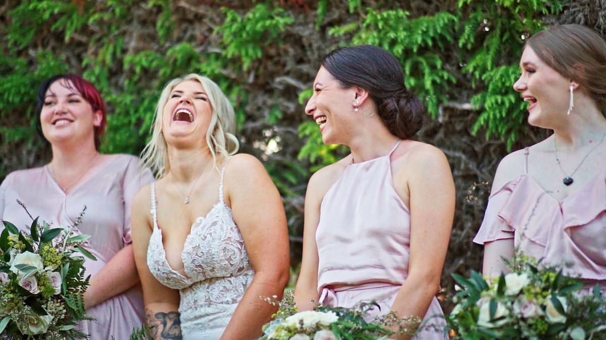 Bride in lace gown laughing joyfully with bridesmaids in blush pink dresses holding bouquets outdoors.