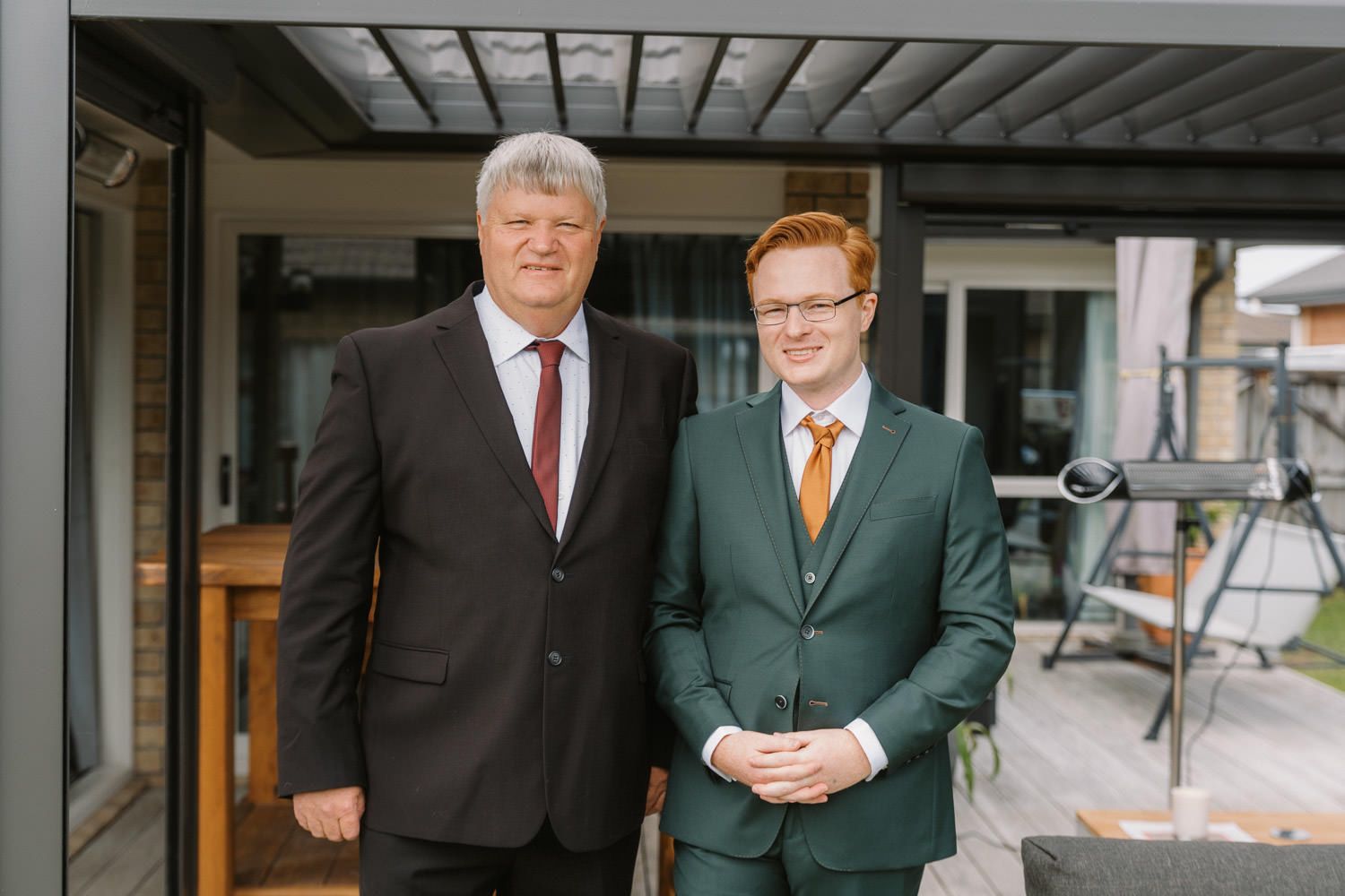 Two men in formal suits smiling together on an outdoor patio, one in dark grey and one in green with an orange tie.