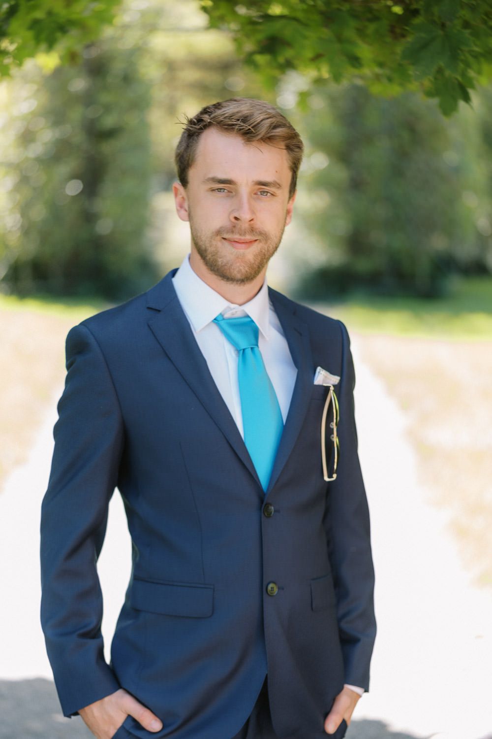Young bearded groom in navy blue suit with turquoise tie and pocket square posing outdoors on sunny day.