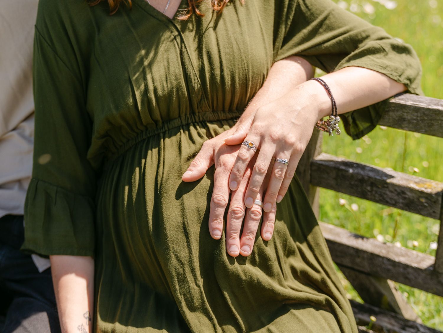 Pregnant woman in green dress with partner's hand on baby bump, standing by wooden fence outdoors.