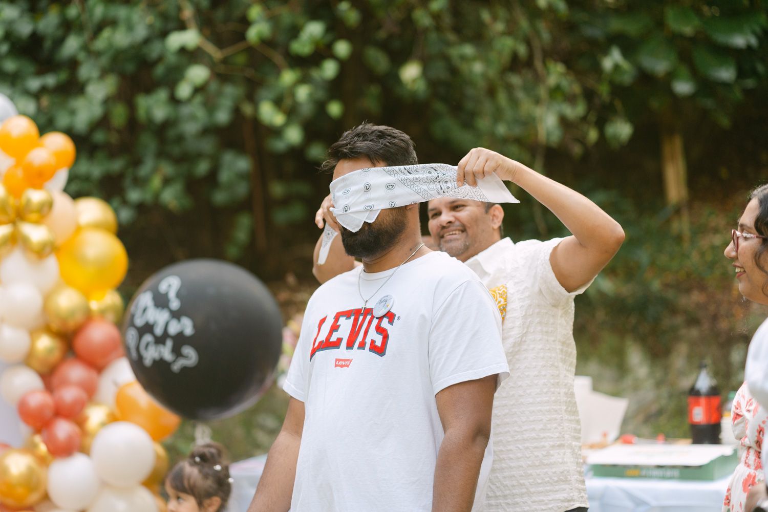 Man blindfolded at outdoor party with balloons, wearing Levis shirt, while friend adjusts blindfold.