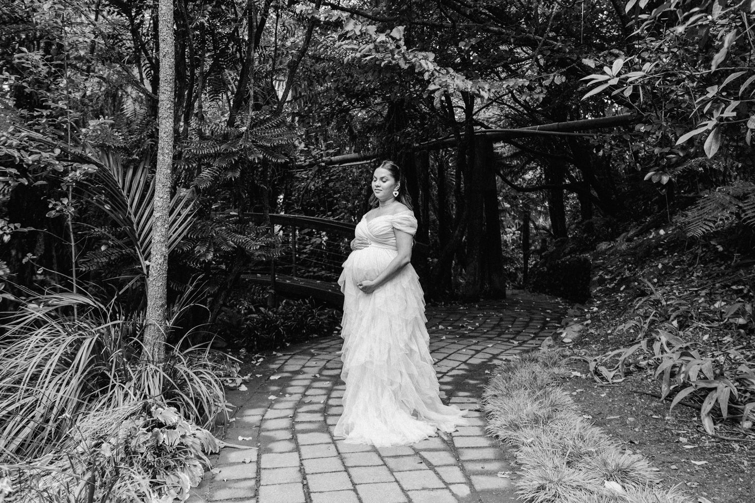 Black and white maternity photo of a pregnant woman in a lace gown standing on a brick path surrounded by lush greenery.