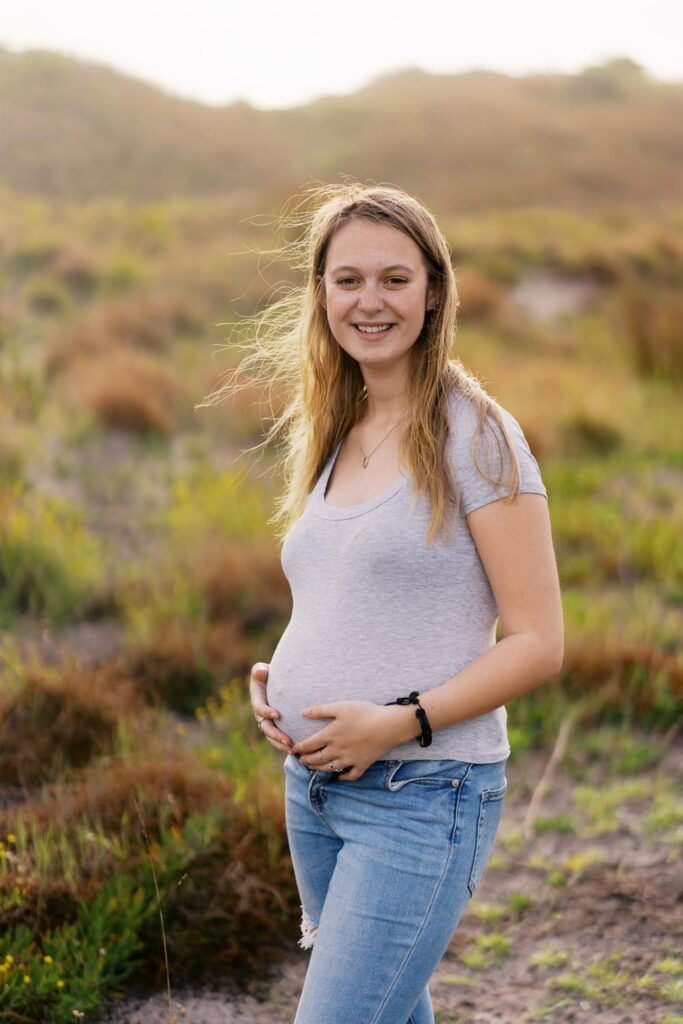 Smiling pregnant woman in gray t-shirt and jeans standing in a sunlit field with hills in the background.