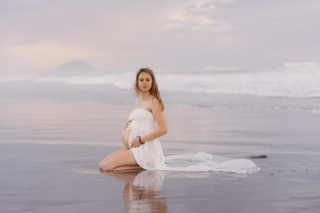 Pregnant woman in white dress sitting on reflective beach at sunset with ocean waves and misty mountains in background.