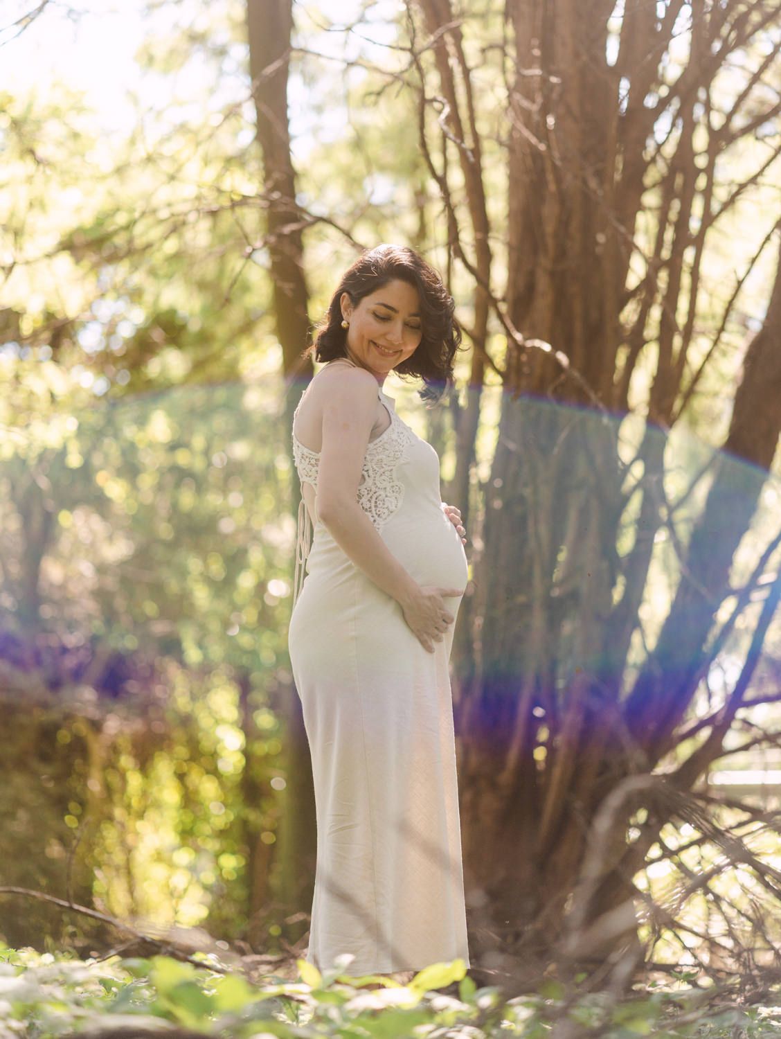 Pregnant woman in white lace gown cradling baby bump outdoors among trees in soft natural light.