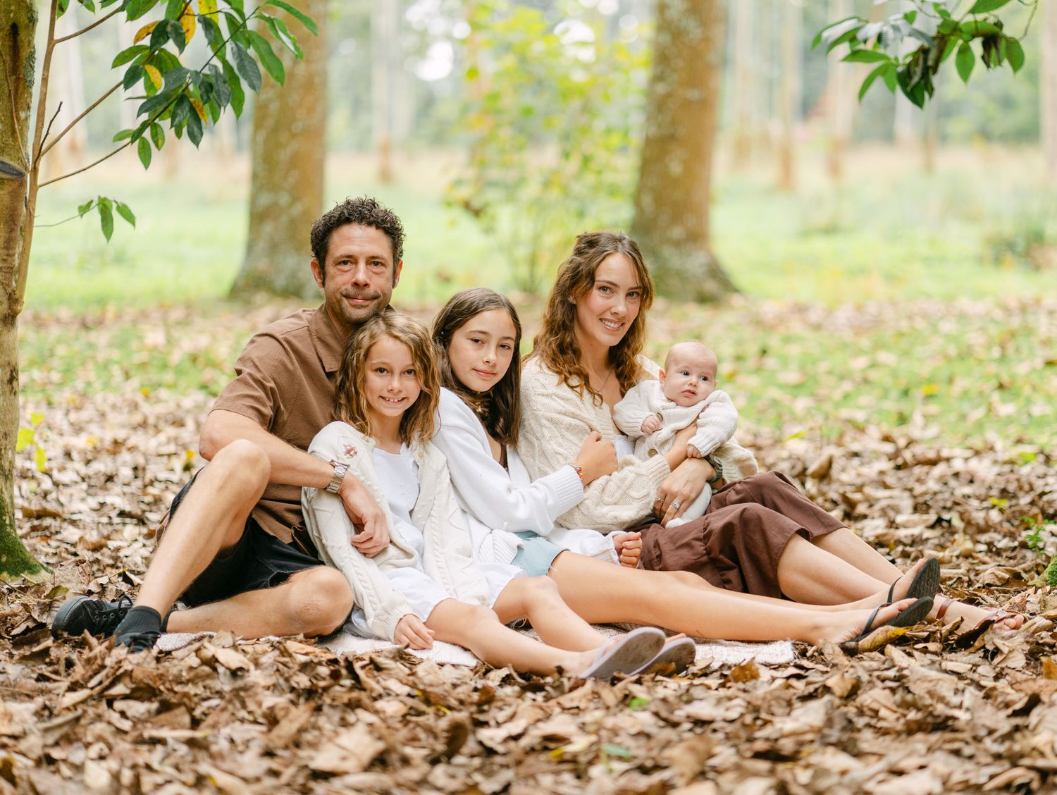 Family of four sitting on autumn leaves in a park, parents with two daughters smiling outdoors.